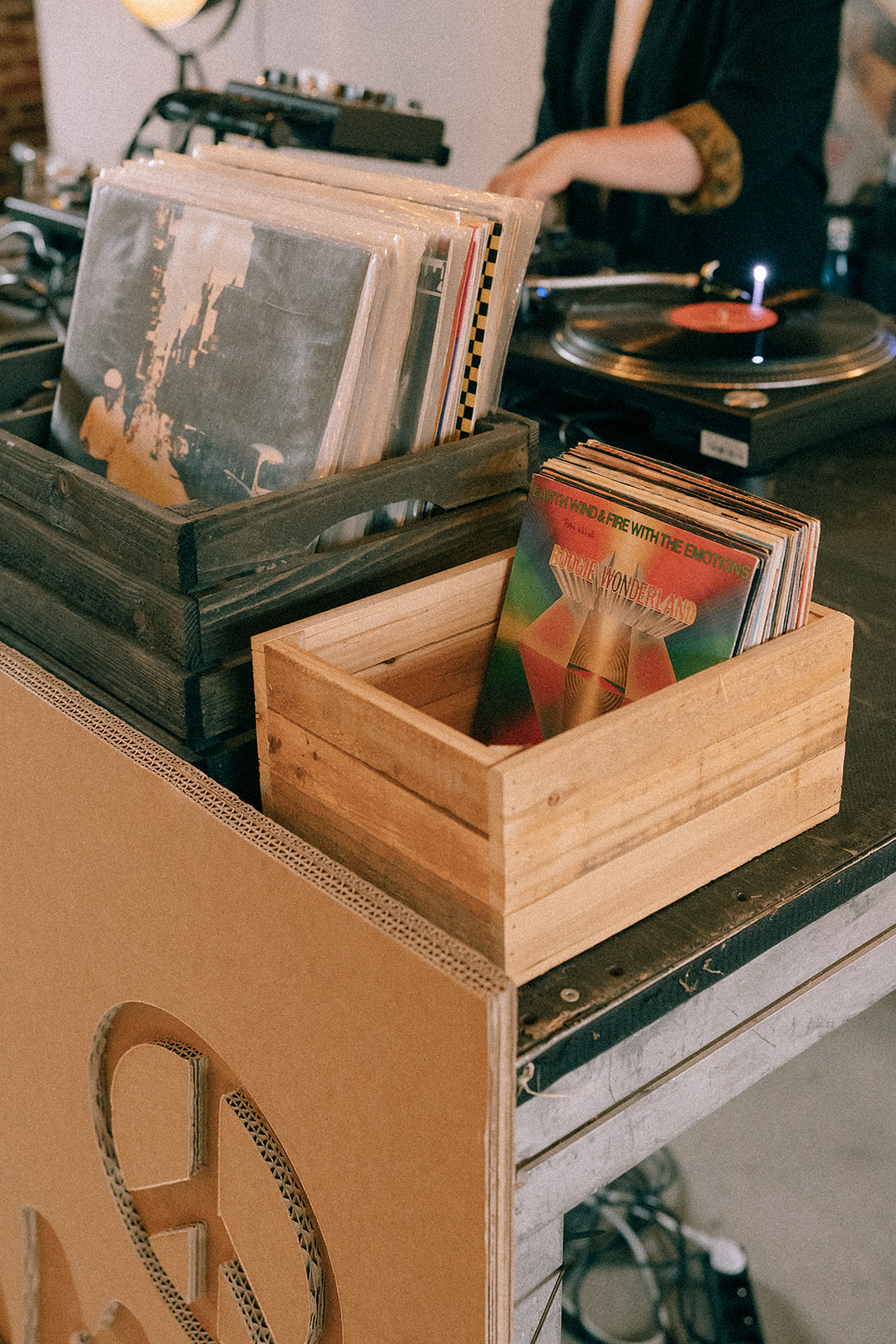 Drewniane skrzynki pełne winyli w kąciku DJ-skim; na wierzchu singiel „Boogie Wonderland” (Earth, Wind & Fire), gramofon w tle.