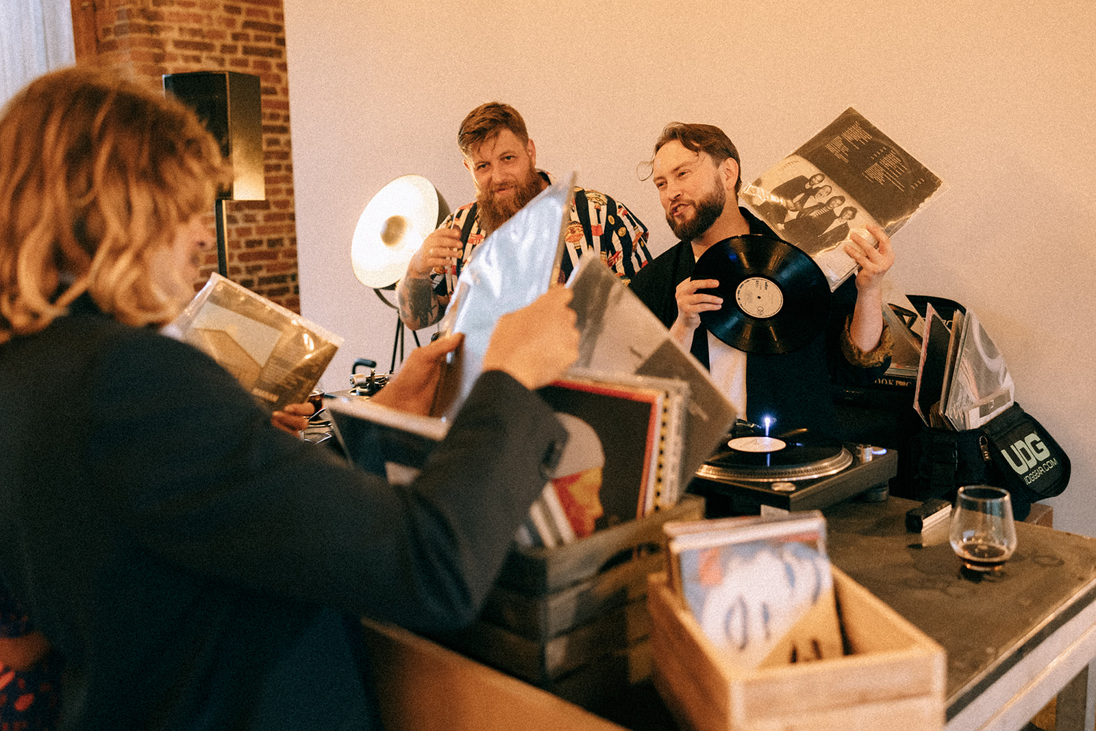 Ruch przy konsolecie: DJ pokazuje czarną płytę, obok osoby wybierają okładki z pełnych skrzynek winyli.