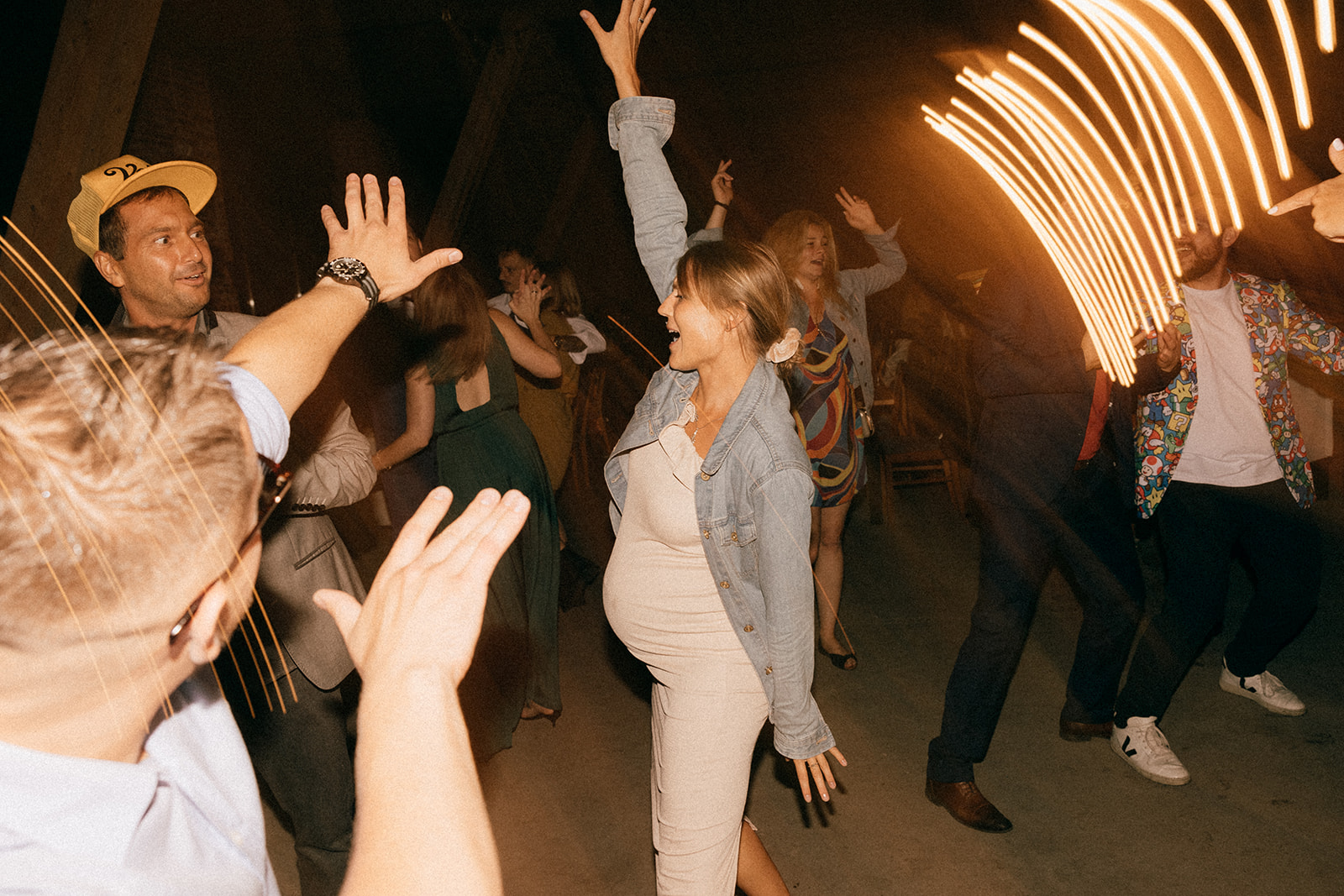 Ludzie tańczą na parkiecie, w centrum kobieta w jasnej sukience z ręką w górze, wokół rozmyte światła i dynamiczny ruch.