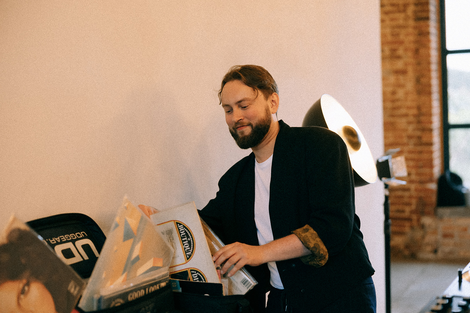 DJ sięga do torby z winylami obok stanowiska; w tle ceglana ściana i lampa studyjna.