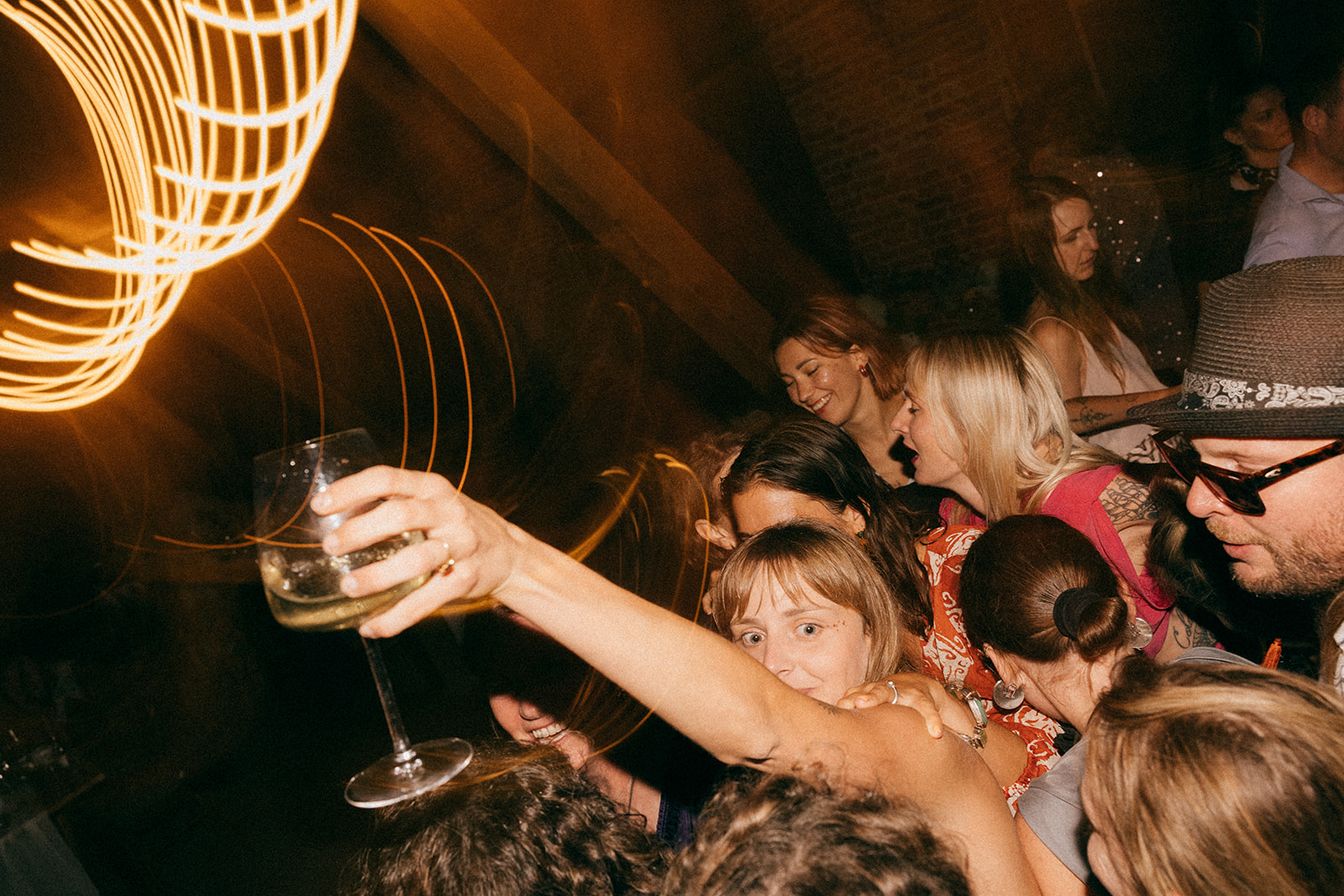 Tłum osób na parkiecie, w centrum kobieta z kieliszkiem wina uniesionym w geście zabawy; energetyczna, nocna atmosfera.