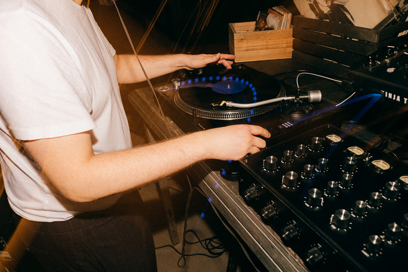 DJ w białej koszulce obsługuje gramofon i mikser, na talerzu wiruje płyta winylowa z niebieskim efektem świetlnym.