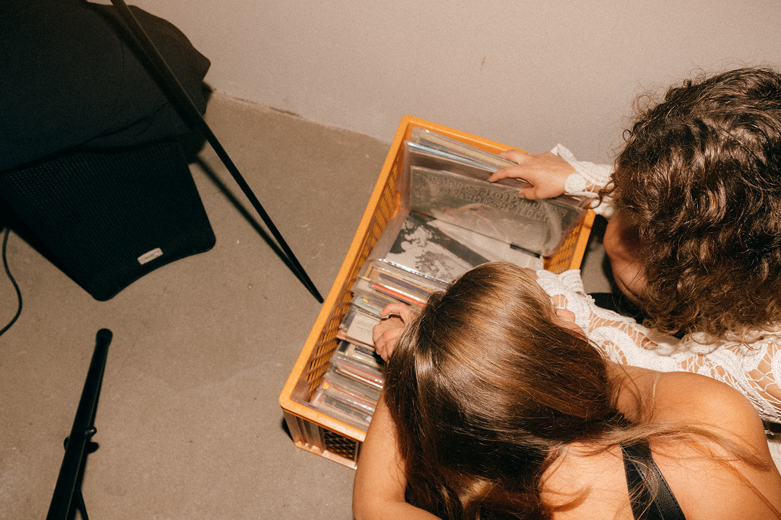 Dwie osoby pochylone nad skrzynką z winylami, przeglądają płyty na podłodze w ciepłym świetle.