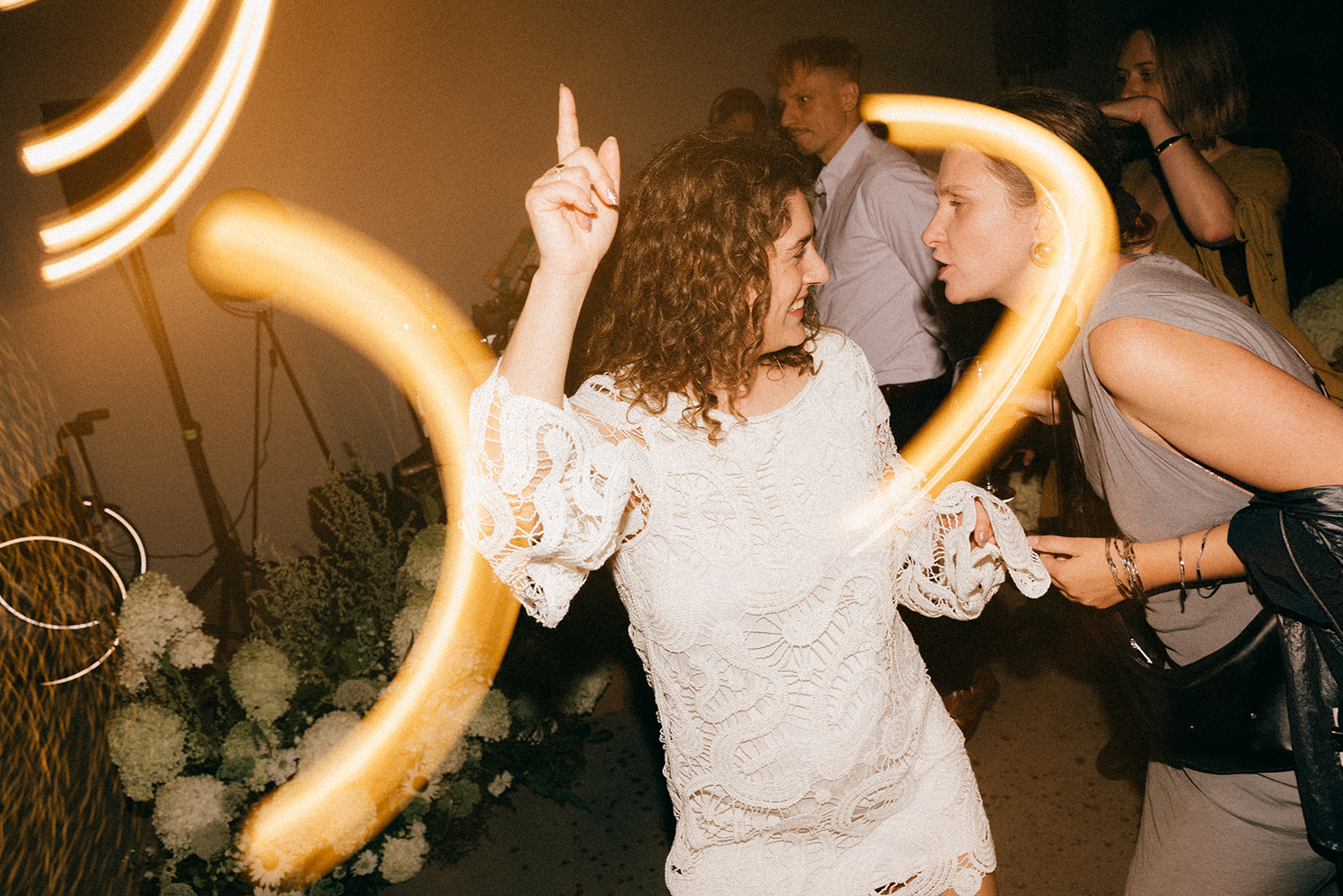 Kobieta w białej koronkowej sukience tańczy, obok druga osoba rozmawia; smugi światła tworzą efekt długiego naświetlenia.