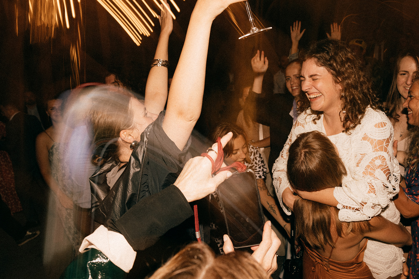 Kobieta w białej koronkowej sukience jest podnoszona i przytulana na parkiecie, wokół uśmiechnięci ludzie z rękami w górze.