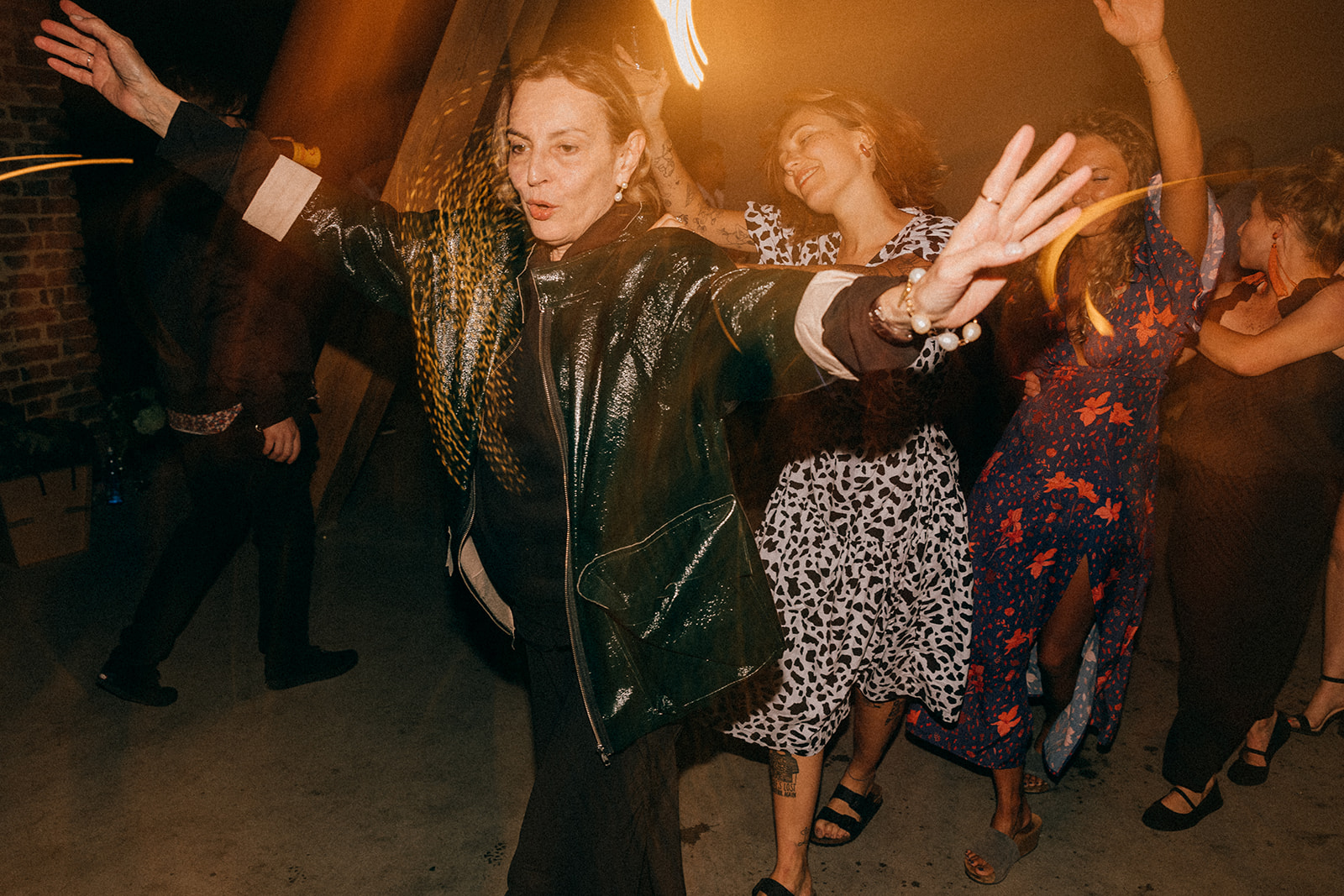 Kobieta w czarnej błyszczącej kurtce tańczy z rozłożonymi rękami, za nią grupa bawiących się osób na parkiecie.