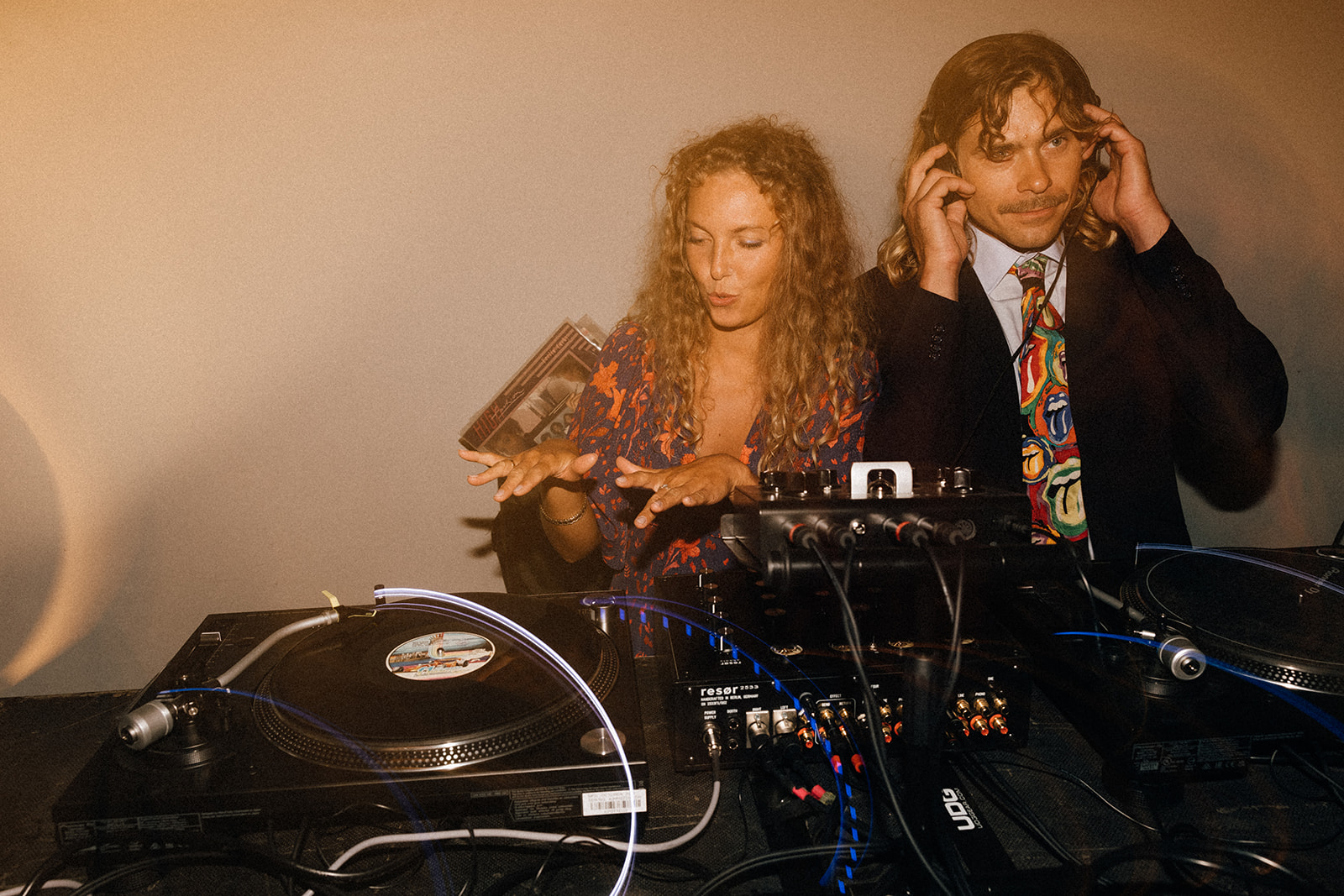 Kobieta udaje miksowanie muzyki, mężczyzna w słuchawkach trzyma je na uchu; stoją przy gramofonach.