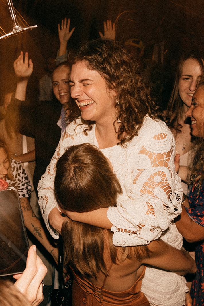 Kobieta w białej koronkowej sukience jest podnoszona i przytulana na parkiecie, wokół uśmiechnięci ludzie z rękami w górze.