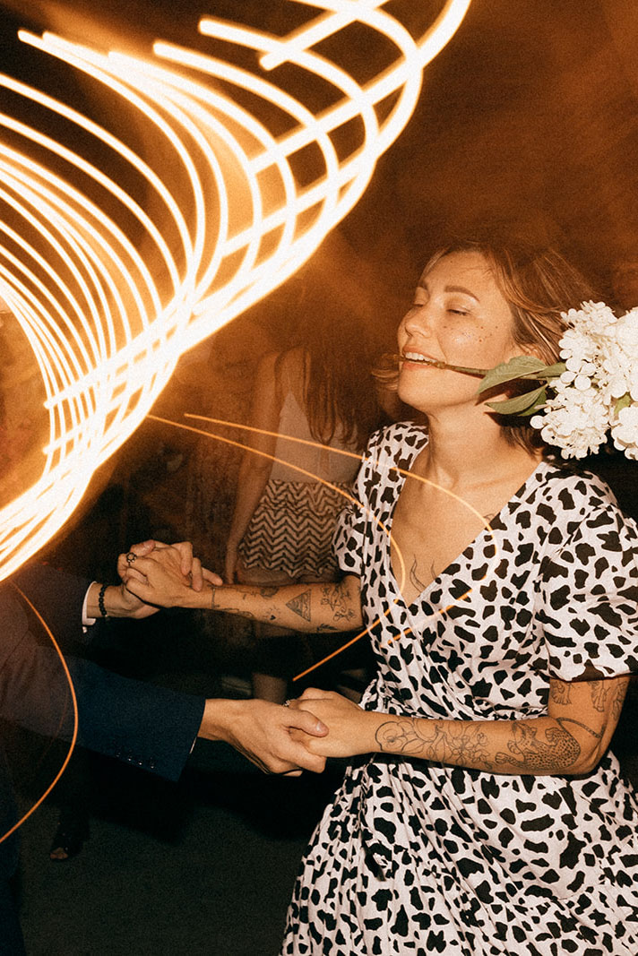 Para tańczy na parkiecie, kobieta trzyma bukiet białych kwiatów w zębach, długie smugi światła w tle.