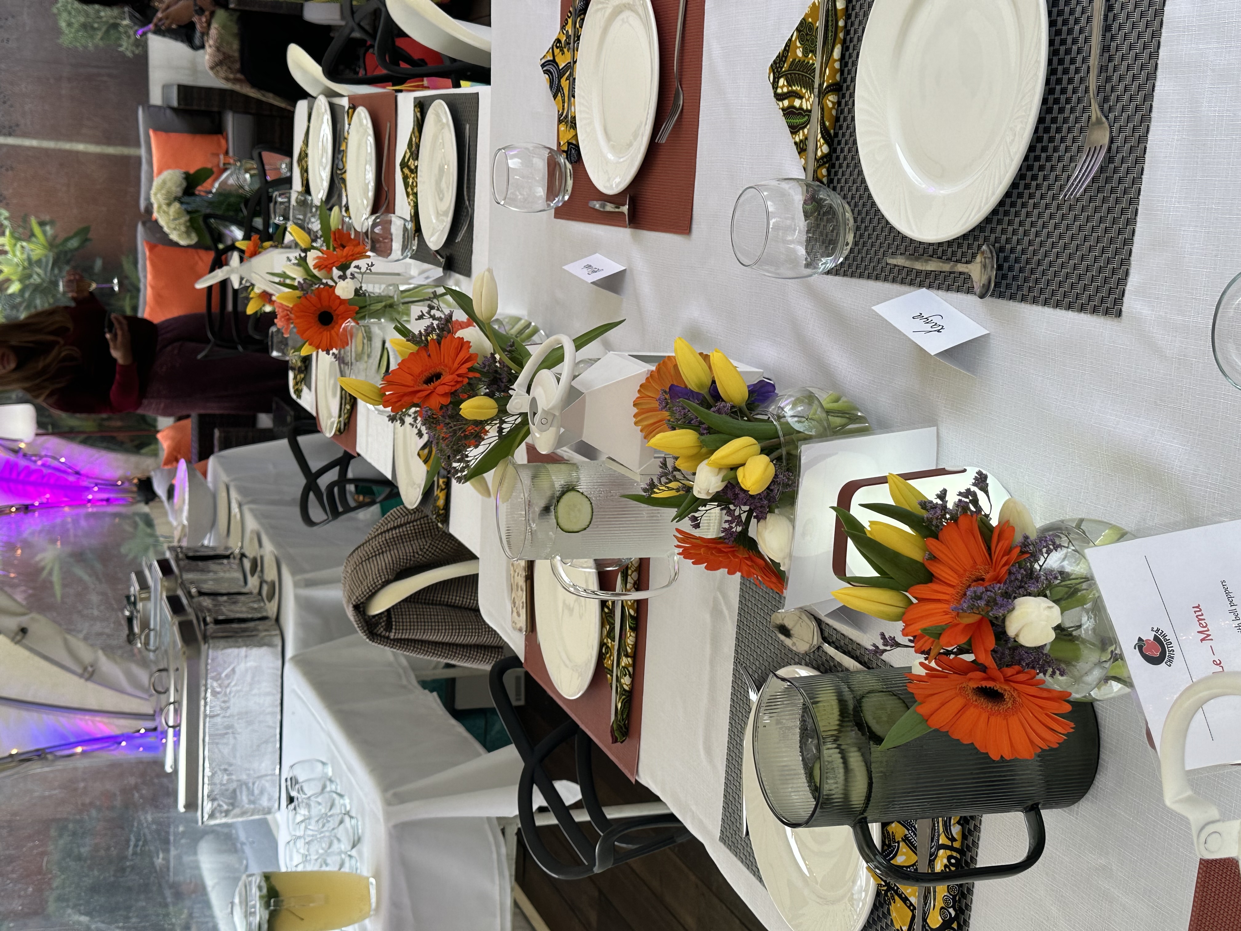 Long dining table set for a meal with white tablecloth, floral centerpieces featuring orange and yellow flowers, pitchers of cucumber water, plates, silverware, and folded patterned napkins, with buffet trays and guests in the background.