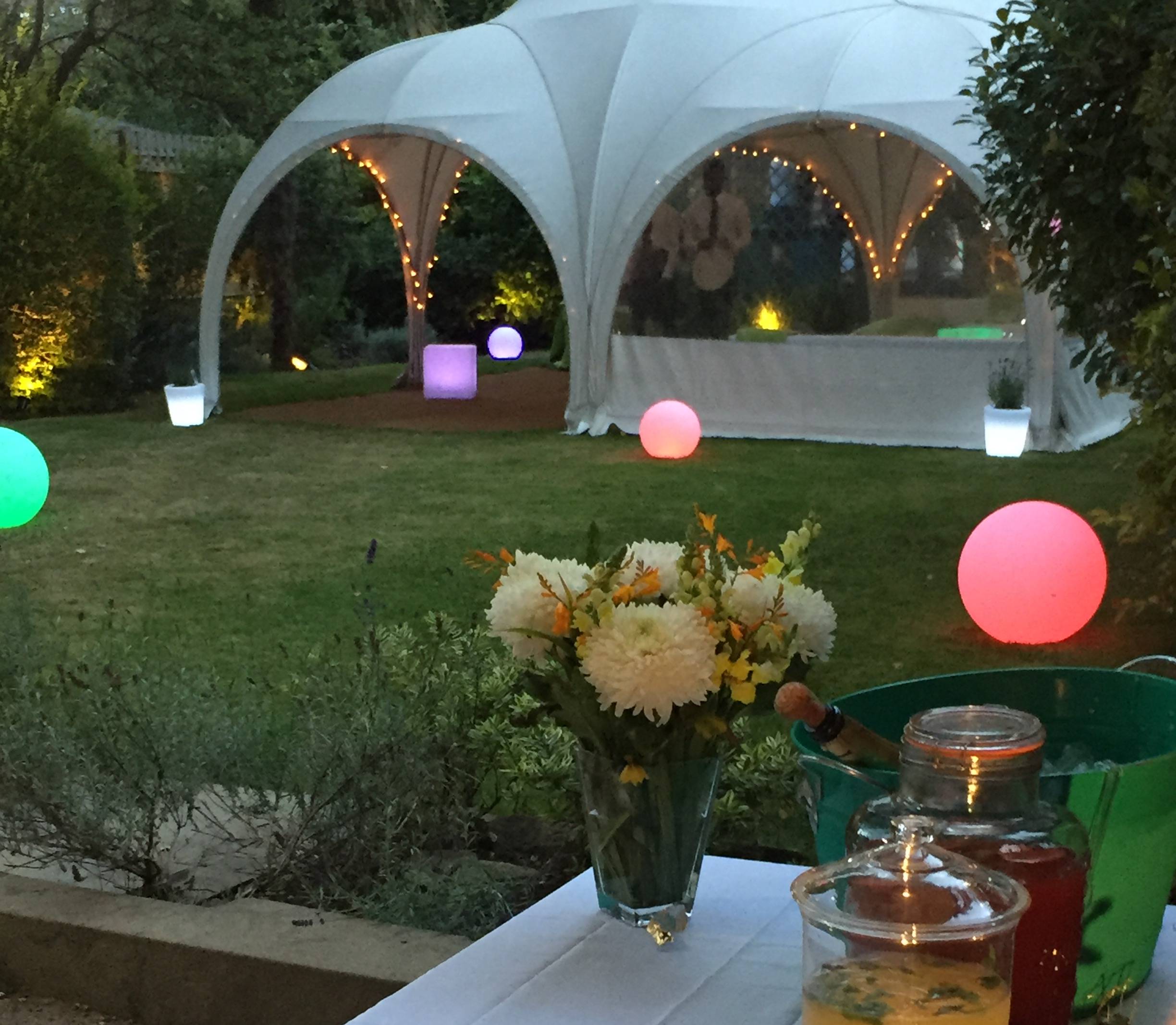 Outdoor evening party setup with a white canopy tent decorated with string lights, glowing colorful spherical lights on grass, a vase of white and yellow flowers on a table, and drinks in glass jars.