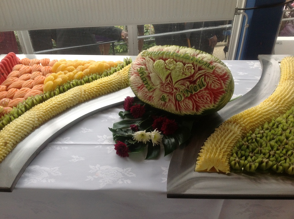 Carved watermelon centerpiece with floral designs and the names 'Shani & O'Neill' surrounded by trays of assorted carved fruits arranged on a table.