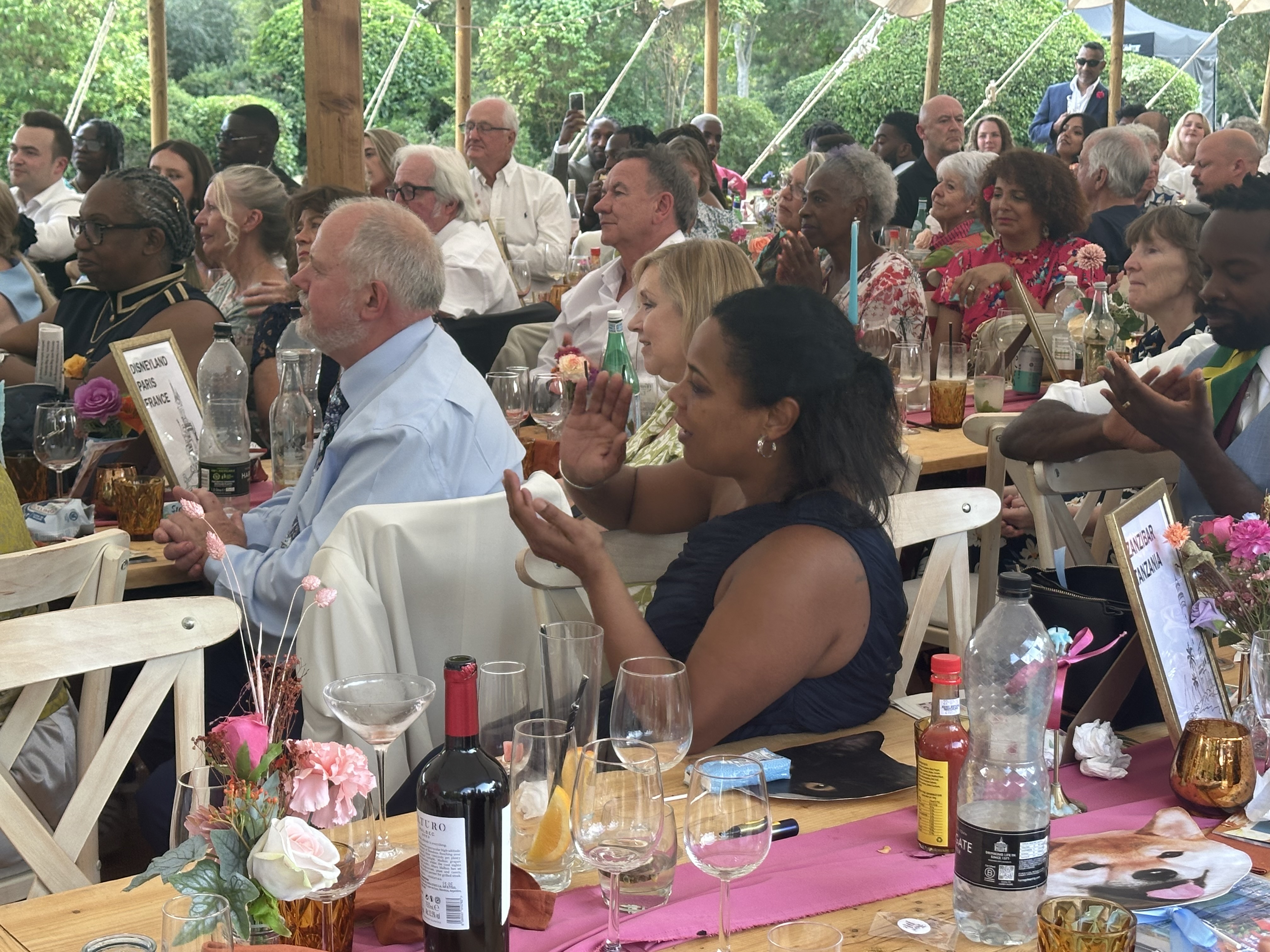 Diverse group of people seated at decorated tables under a tent, attentively clapping during an event.