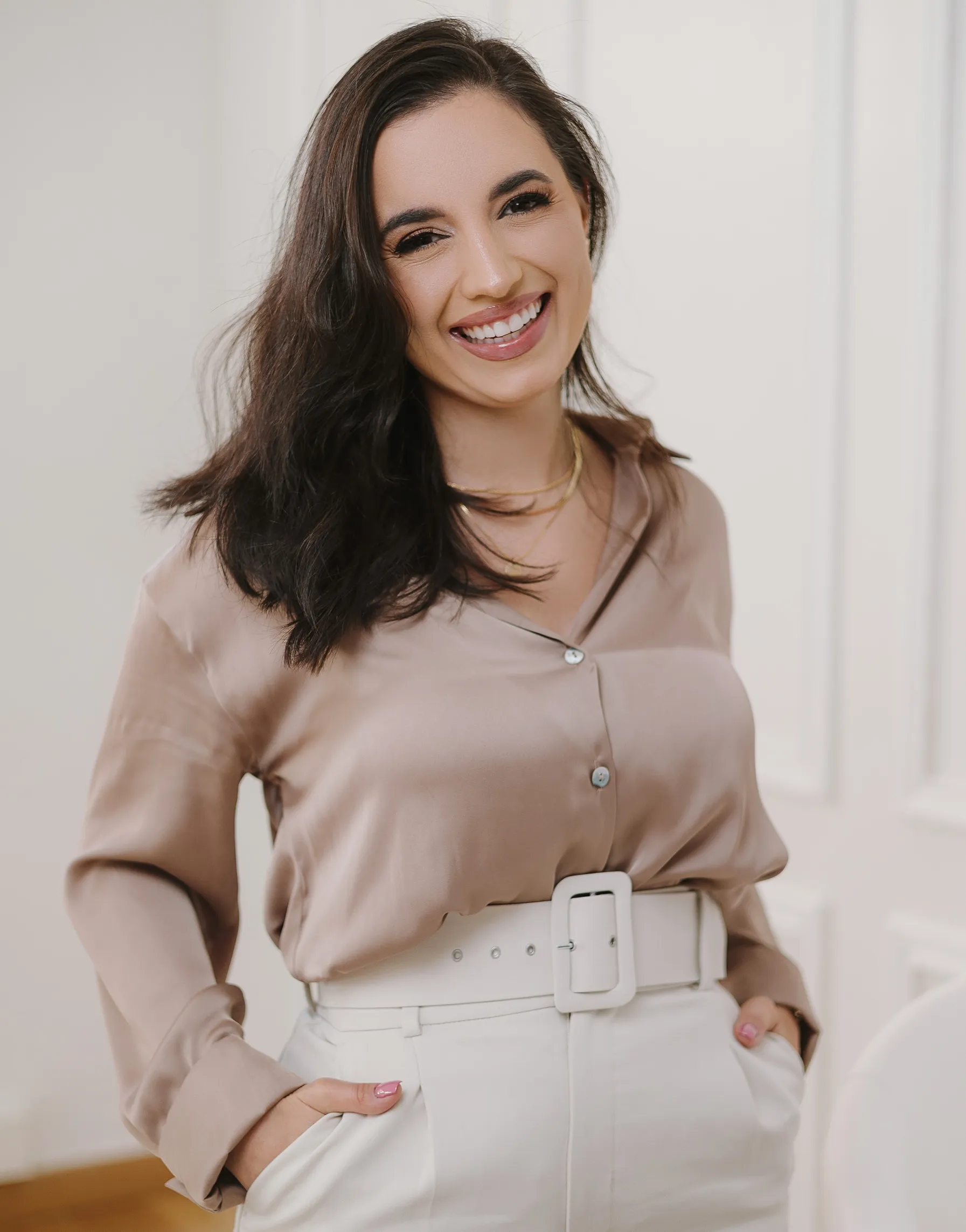 Mulher sorridente de cabelos castanhos escuros, camisa bege e calças brancas com cinto largo.