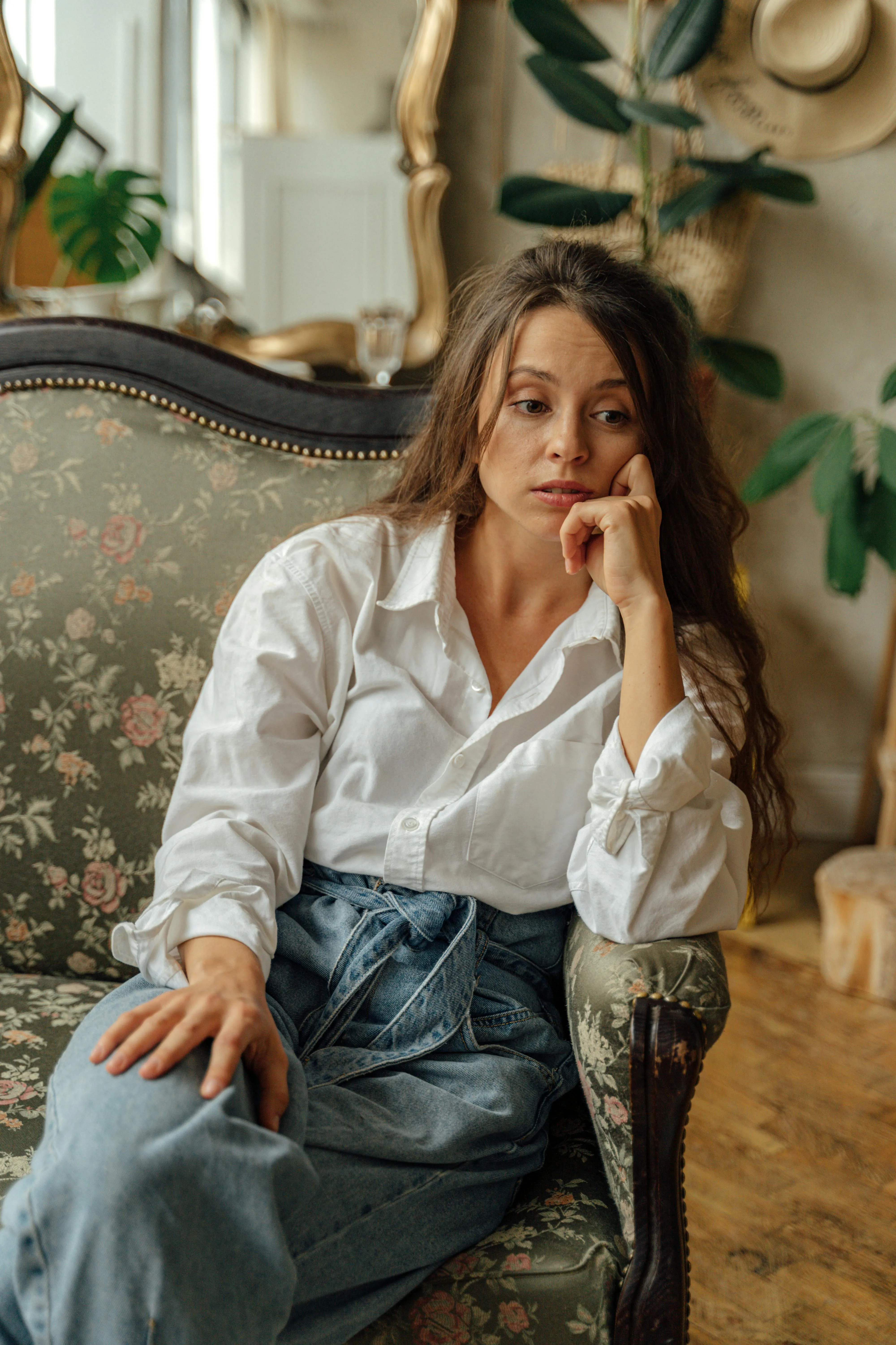 Mulher pensativa sentada numa poltrona floral com camisa branca e jeans azuis.