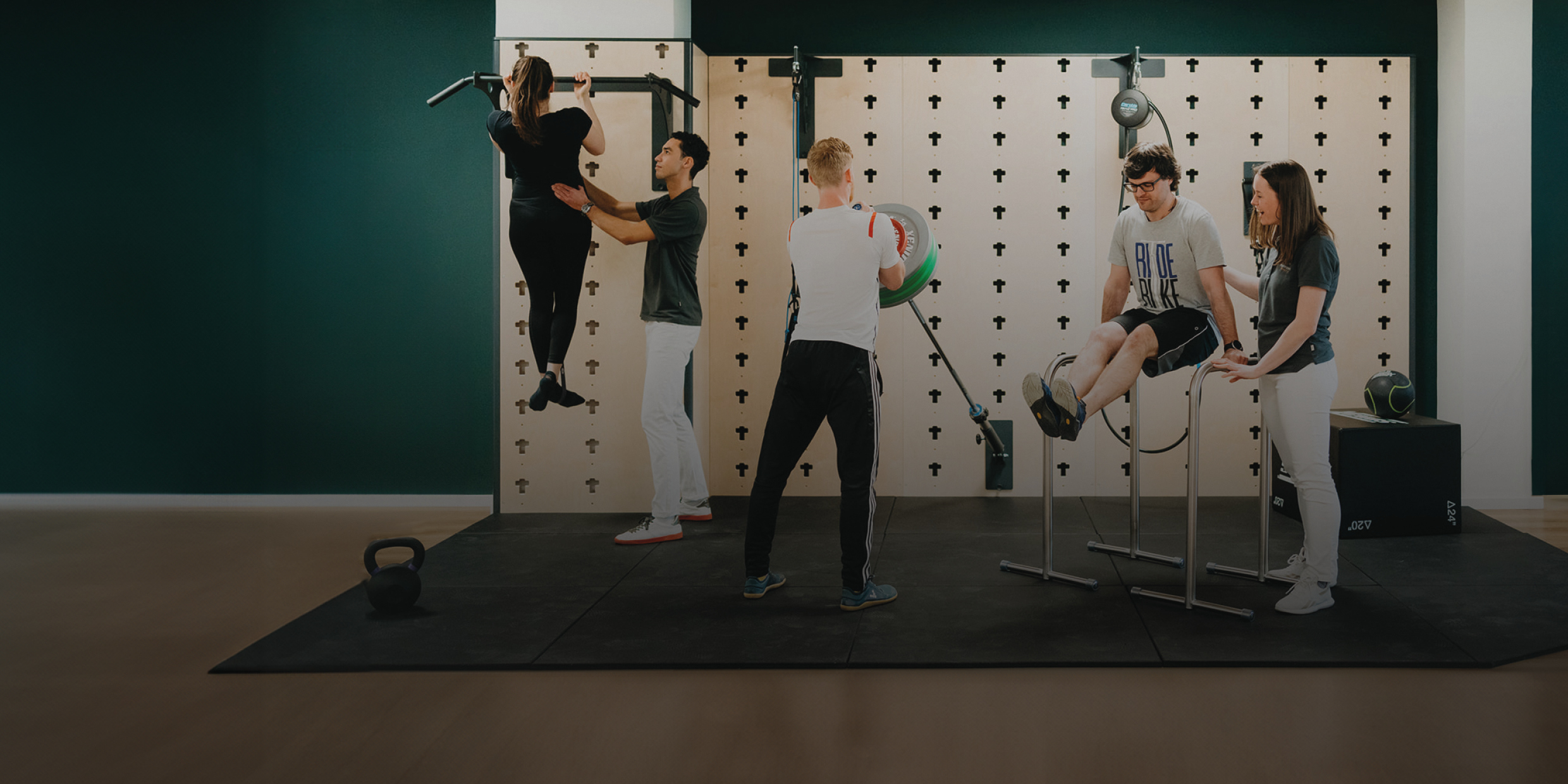 Vier Personen üben verschiedene Fitnessübungen in einem modernen Trainingsraum mit Kettlebell und Hantelwand.