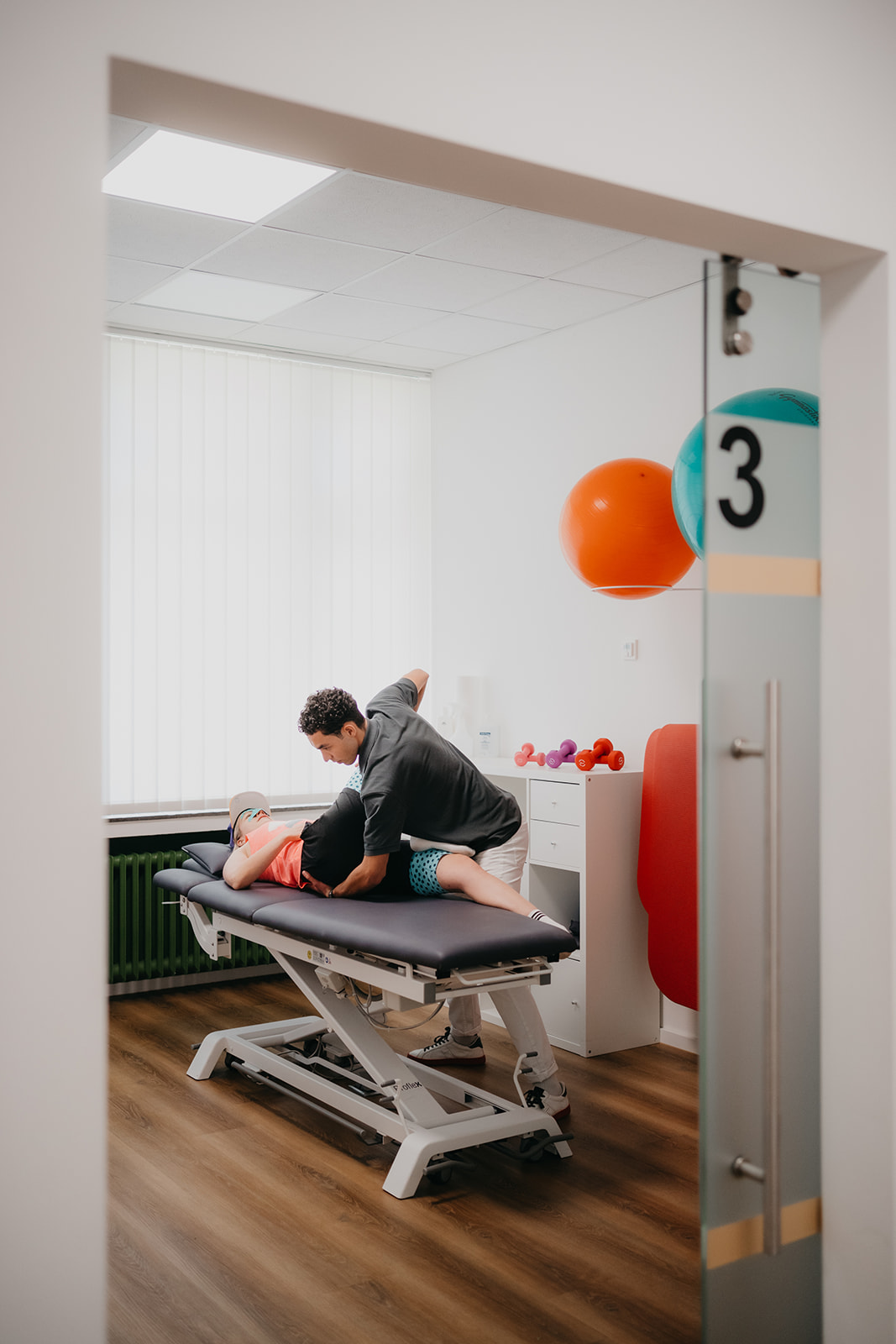 Ein Physiotherapeut behandelt einen Patienten auf einer Behandlungsliege in einem hellen Raum mit Gymnastikbällen und Hanteln.