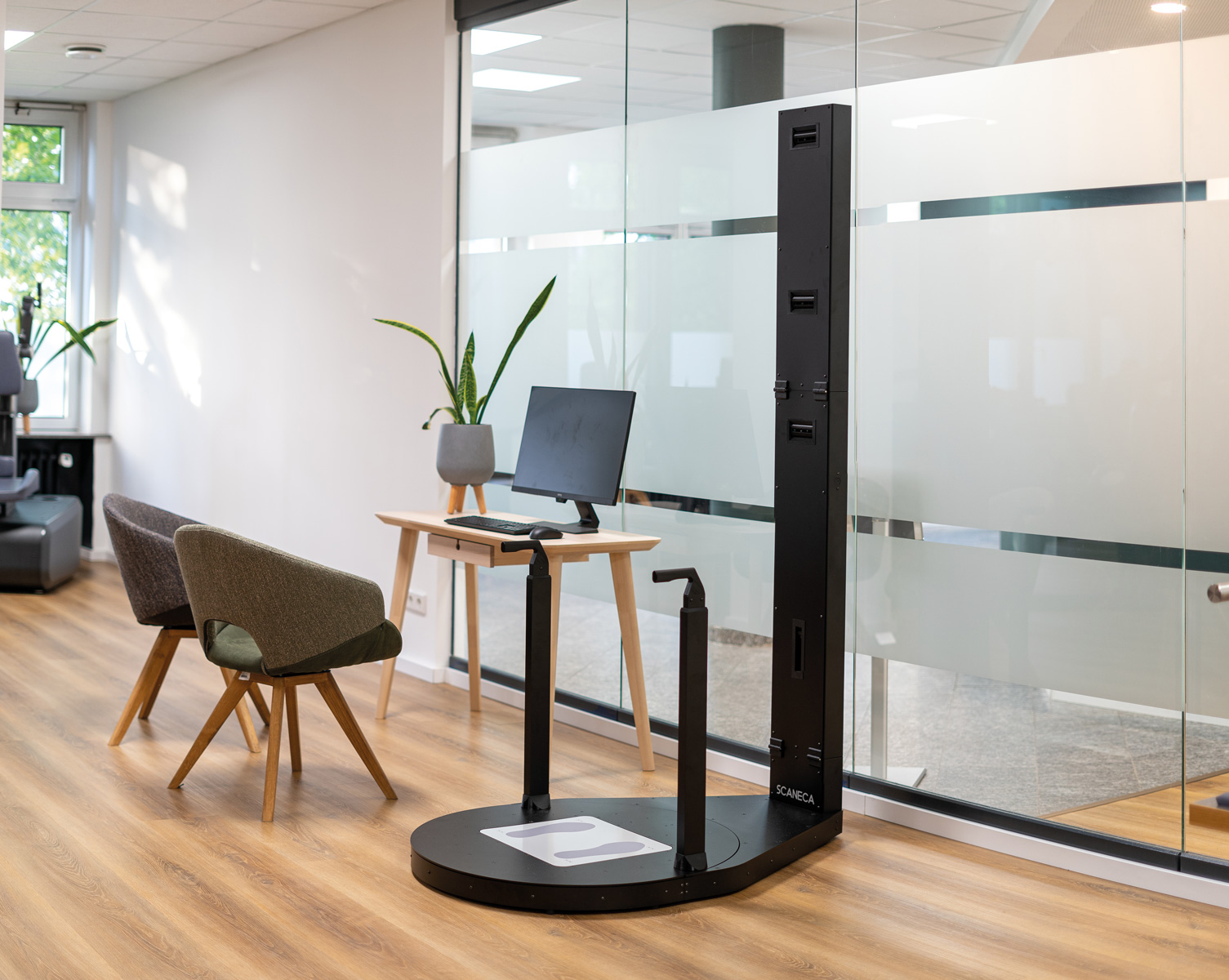 Modernes Körperanalysegerät mit schwarzer Basis und Bildschirm in hellem Raum mit zwei Stühlen und Holzfußboden.