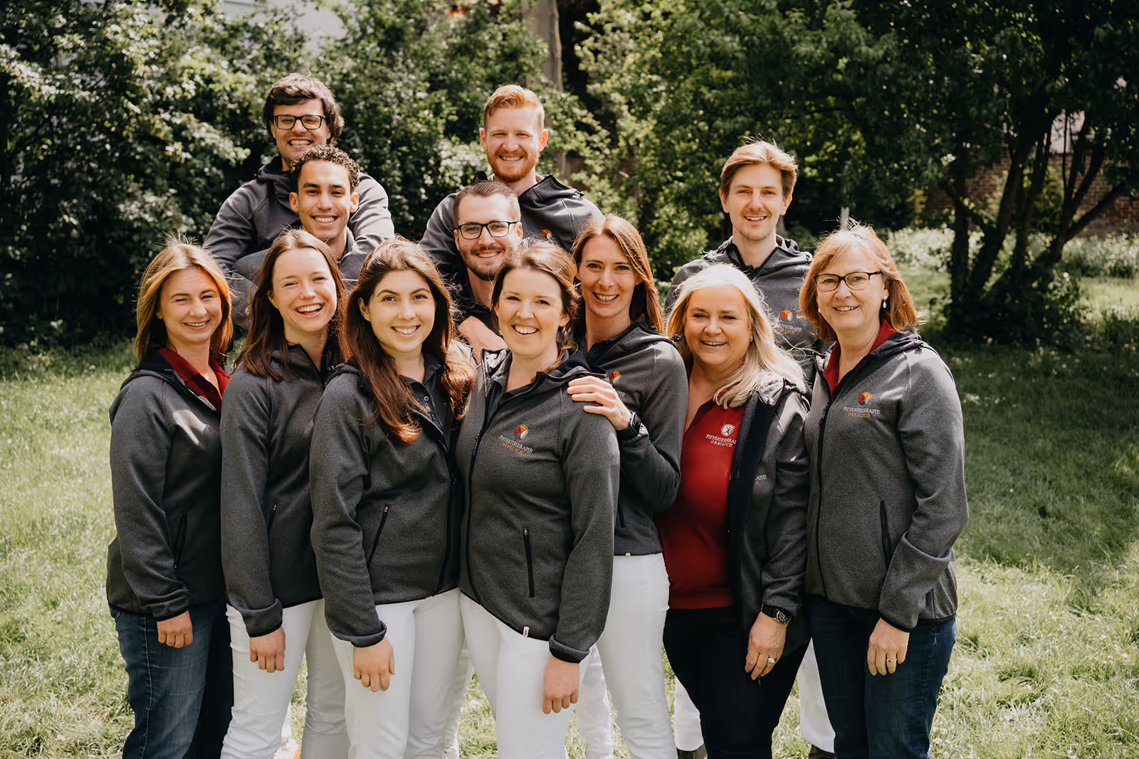 Gruppe von elf lächelnden Personen in grauen Jacken und weißen Hosen, die draußen in einem grünen Park stehen.