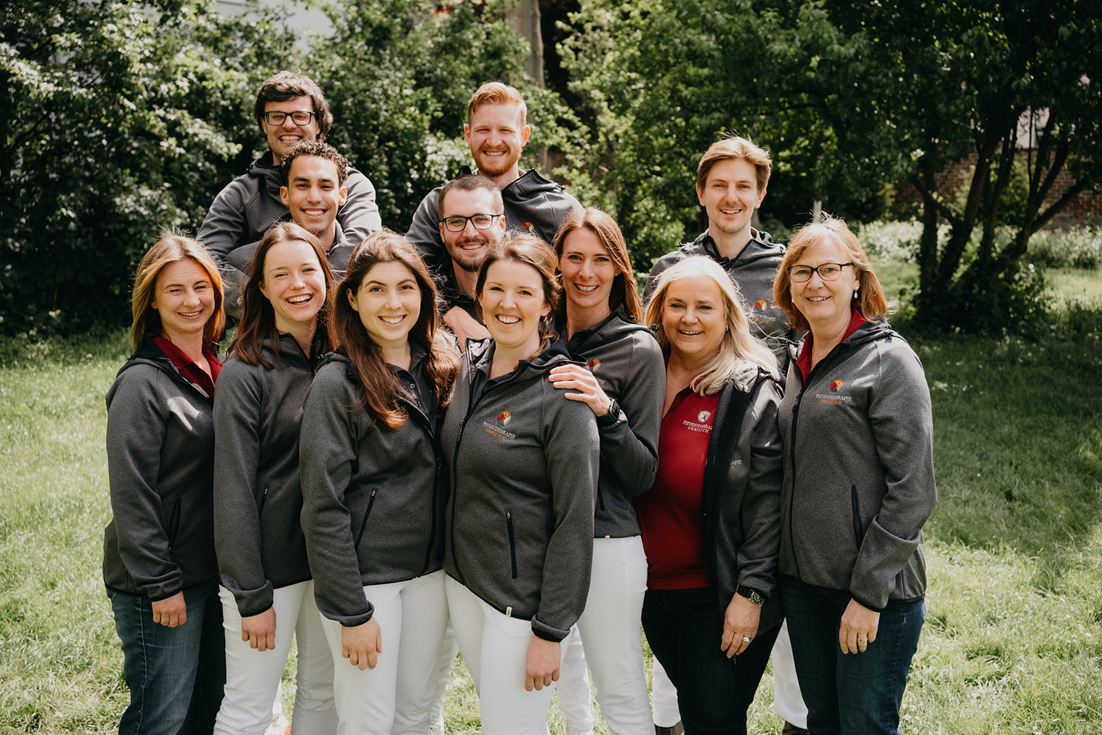 Gruppe von elf lächelnden Personen in grauen Jacken und weißen Hosen, die draußen in einem grünen Park stehen.