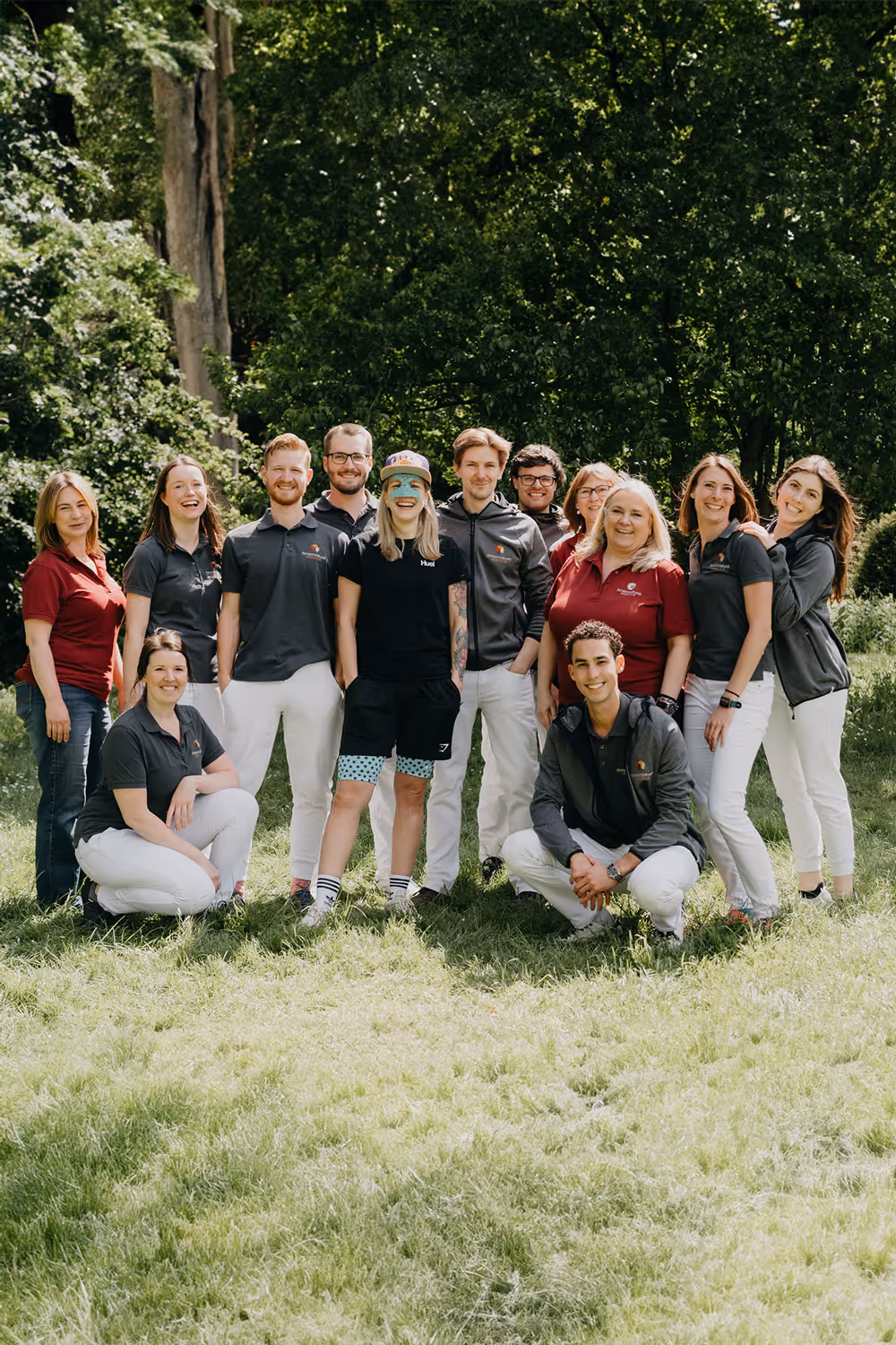 Zwölf Personen, einige stehend, einige hockend, lachend und entspannt im Freien vor Bäumen in Freizeit- und Arbeitskleidung.