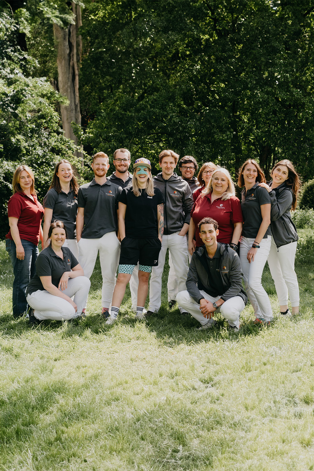 Zwölf Personen, einige stehend, einige hockend, lachend und entspannt im Freien vor Bäumen in Freizeit- und Arbeitskleidung.