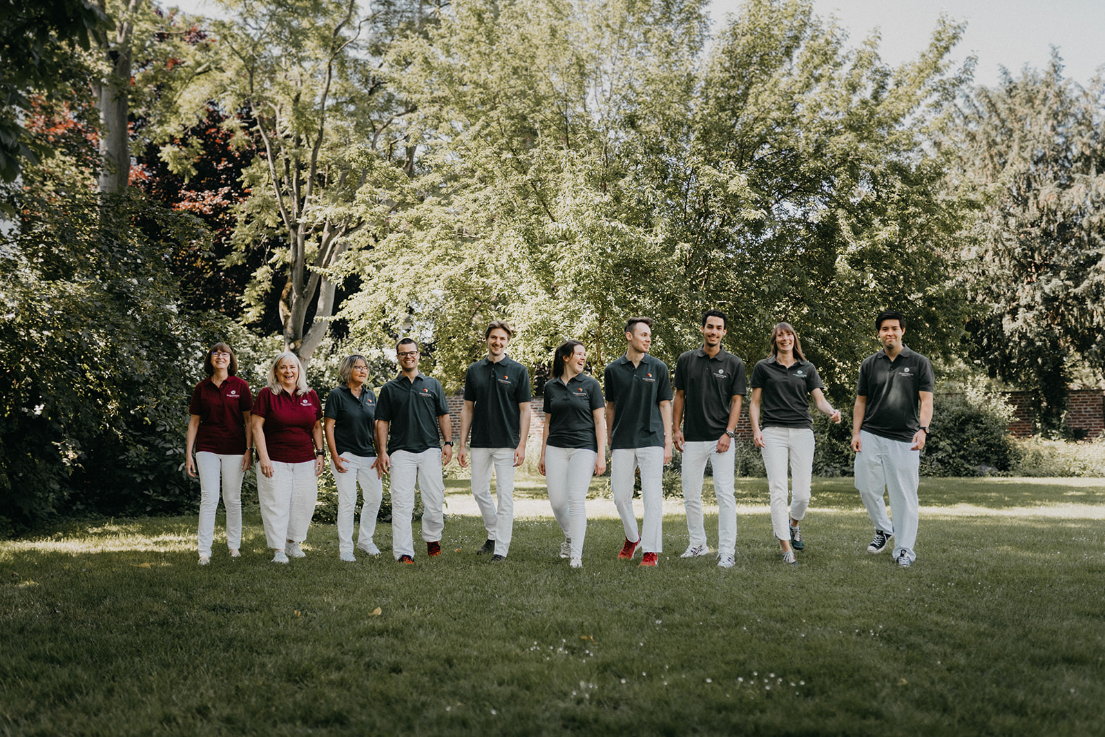 Zehn Personen in weißen Hosen gehen bei sonnigem Wetter auf einer Wiese vor grünen Bäumen, tragen dunkelrote und schwarze Poloshirts.