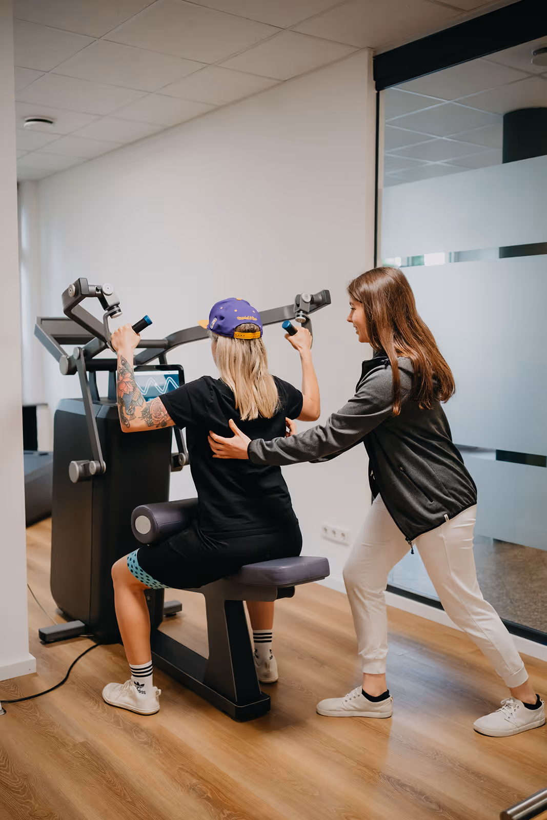 Eine Frau mit blonden Haaren und Tattoo trainiert an einem Fitnessgerät, während eine Trainerin sie von hinten anleitet.