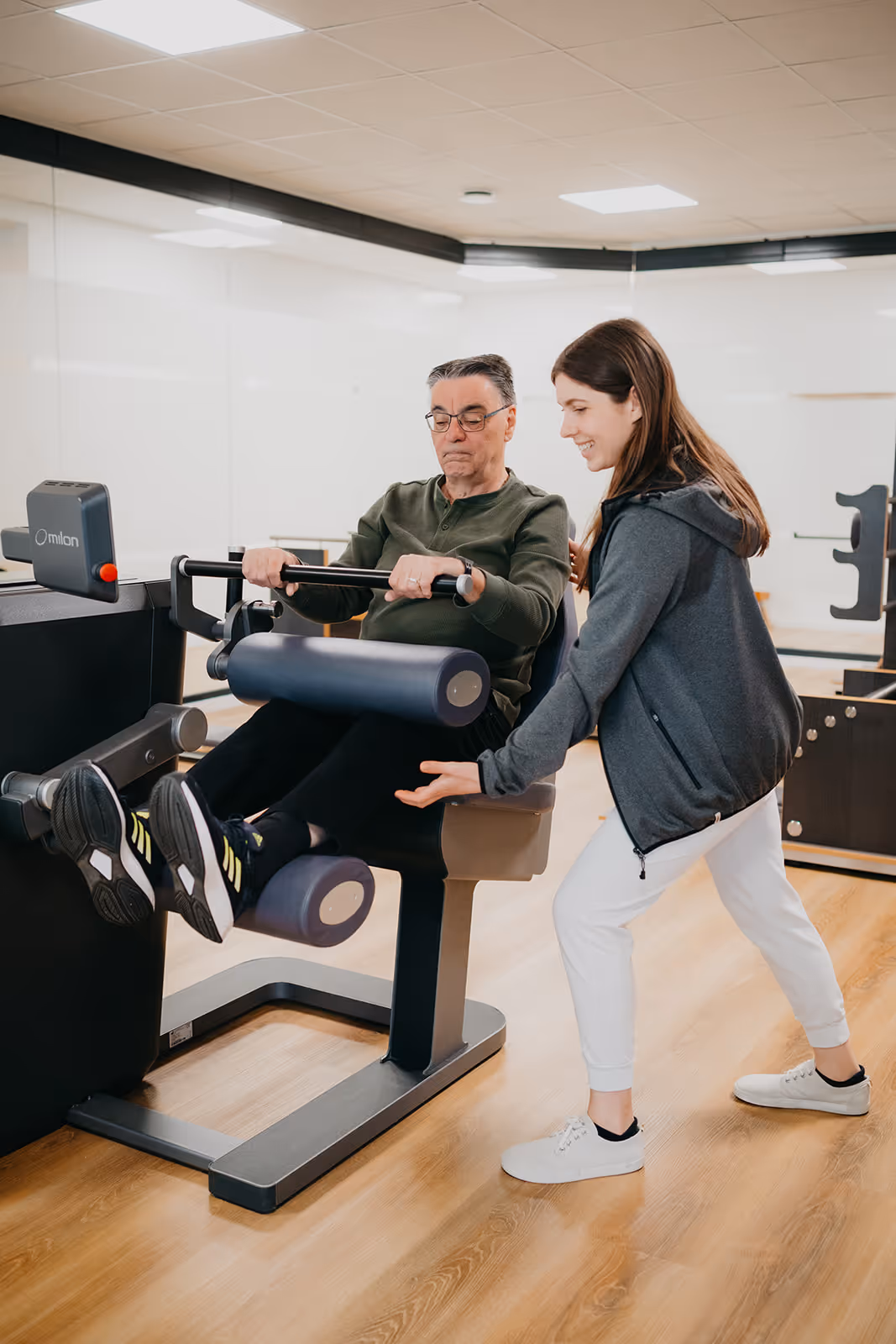 Älterer Mann trainiert an Fitnessgerät zur Beinübung, betreut von junger weiblicher Physiotherapeutin in heller Trainingsumgebung.