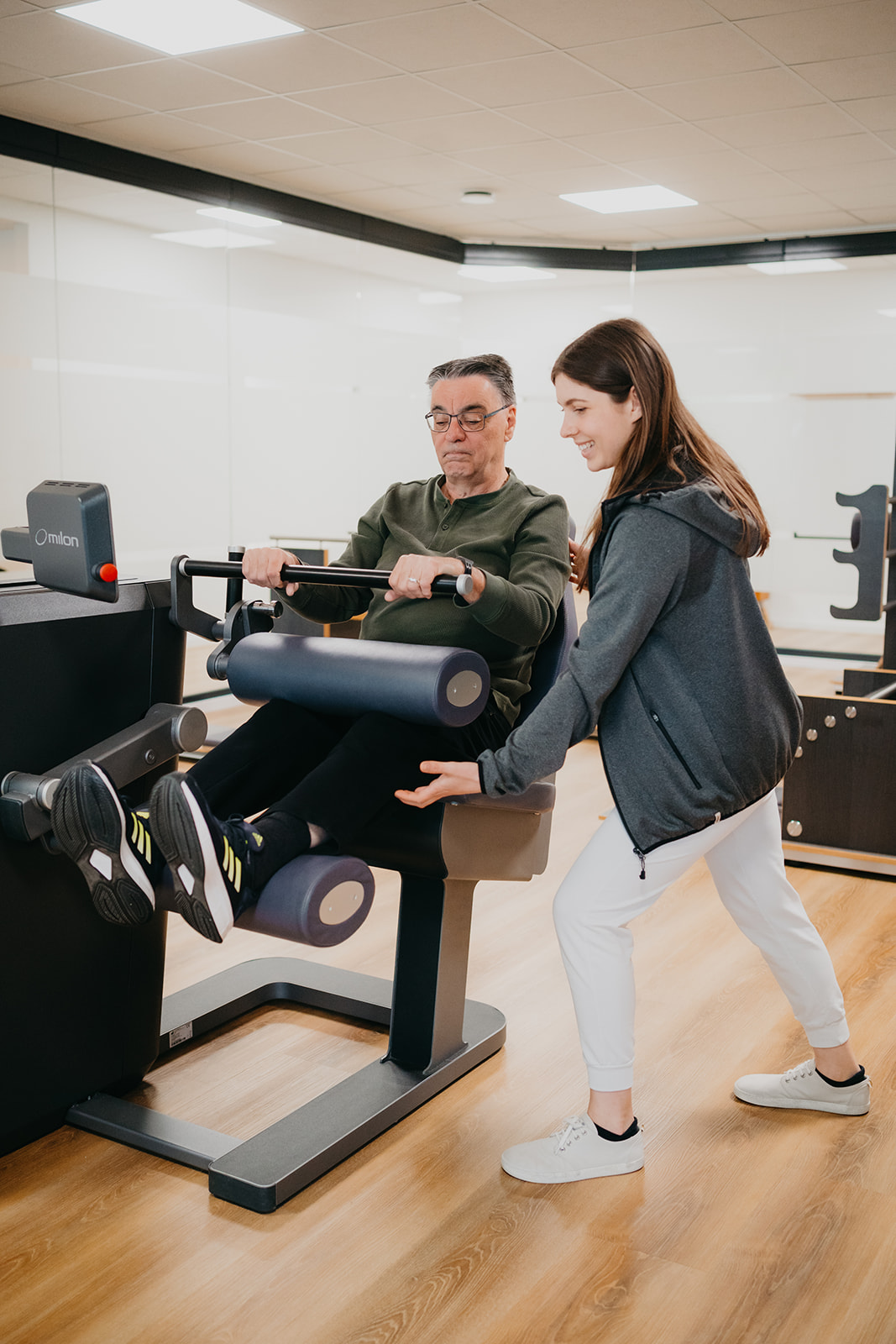 Älterer Mann trainiert an Fitnessgerät zur Beinübung, betreut von junger weiblicher Physiotherapeutin in heller Trainingsumgebung.