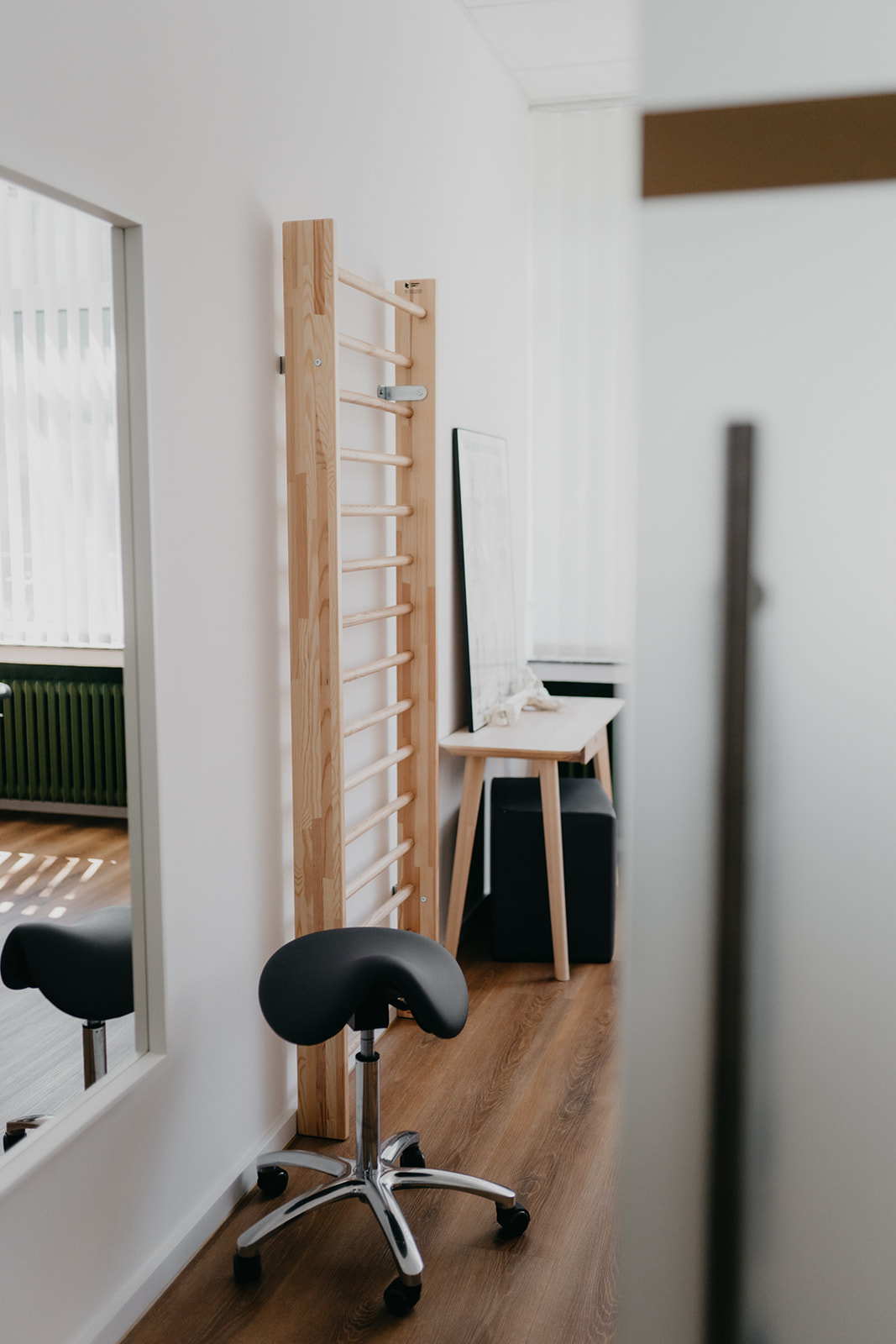 Moderner Behandlungsraum mit Rollhocker, Holz-Kletterwand an der Wand, großem Spiegel und einem Tisch mit schwarzem Hocker.
