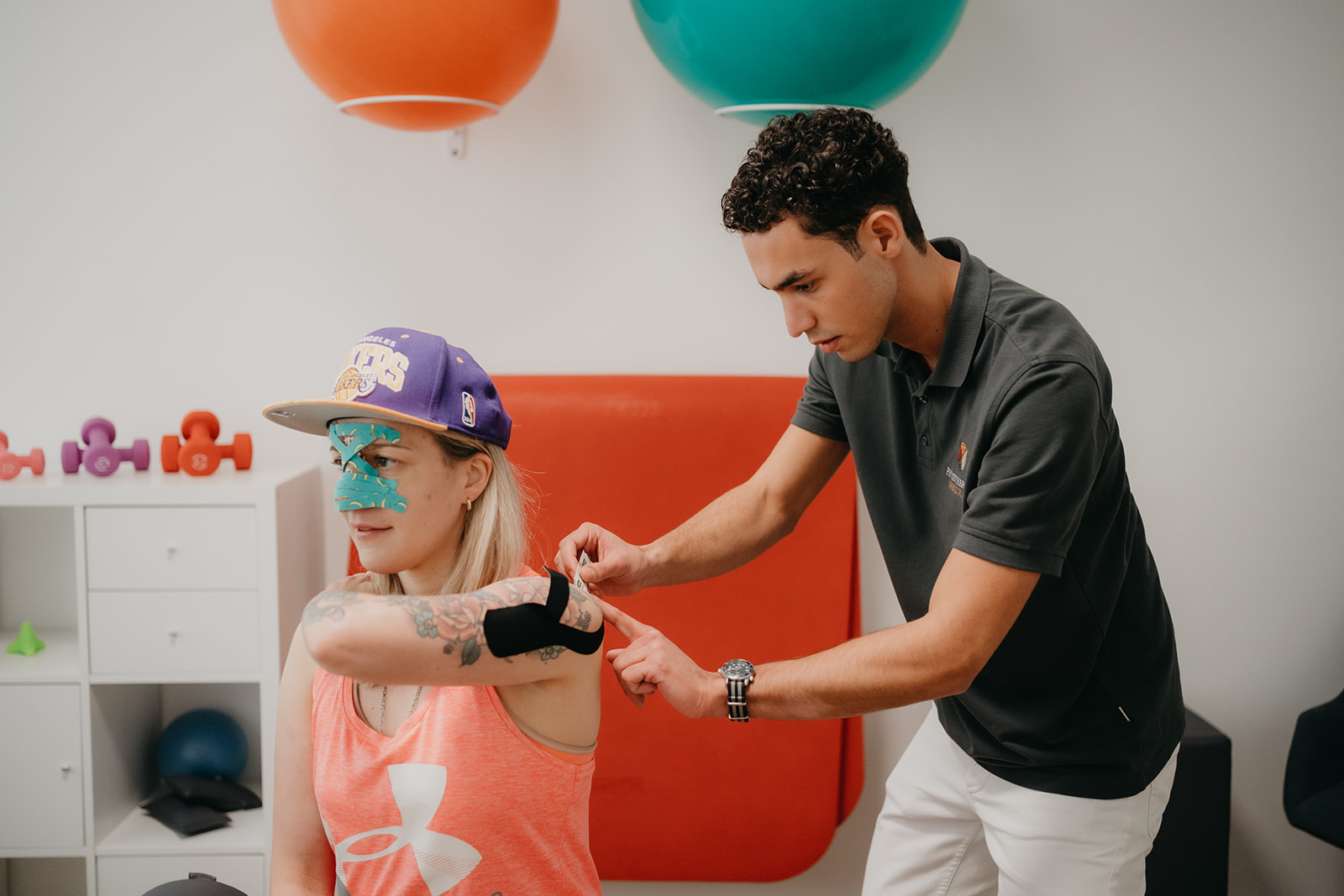 Eine Frau mit Sportkleidung und Tattoos erhält von einem Mann eine therapeutische Kinesiologie-Tape-Anwendung am Oberarm in einer Trainingsumgebung.