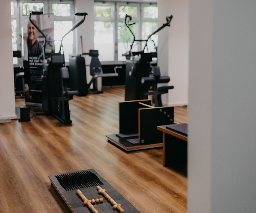 Innenansicht eines Fitnessstudios mit Holzboden, Trainingsgeräten und großen Fenstern im Hintergrund.