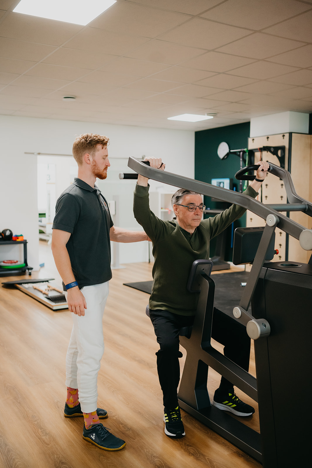 Ein älterer Mann trainiert an einem Kraftgerät, betreut von einem jüngeren Trainer in einem Fitnessstudio.