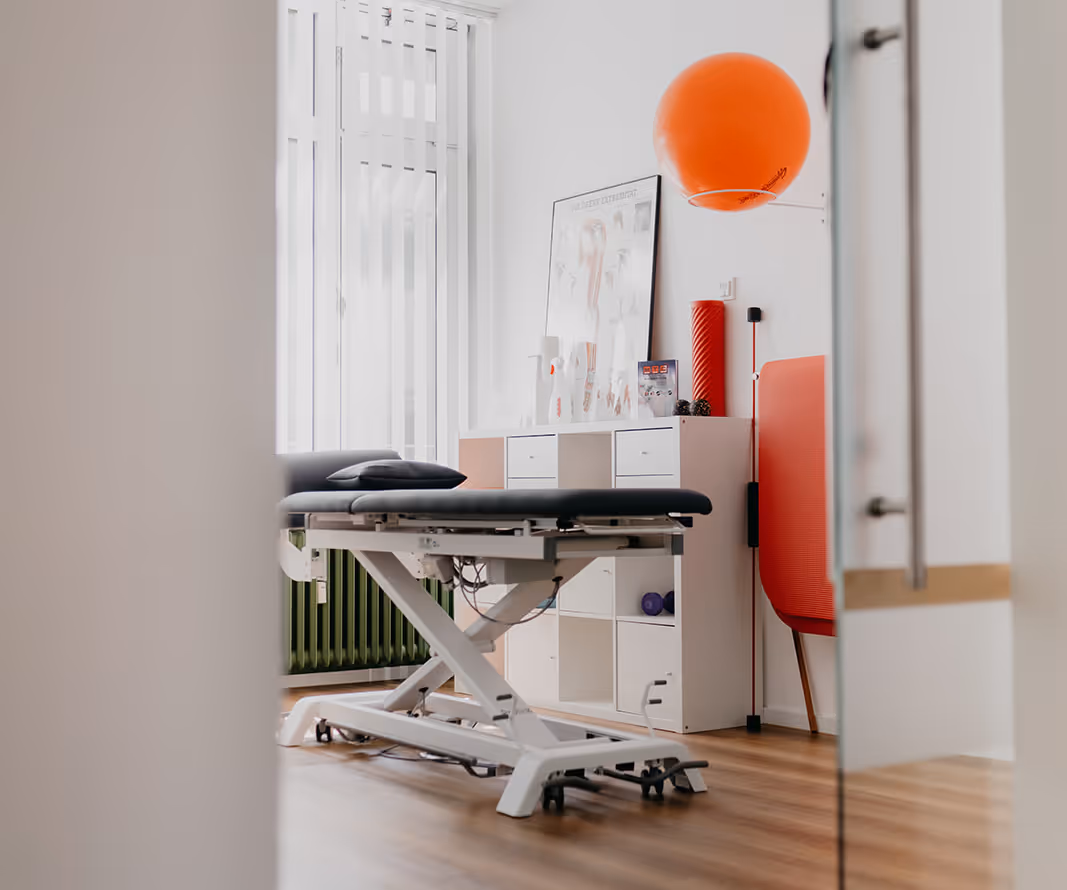 Behandlungsraum einer Physiotherapiepraxis mit verstellbarer Liege, orangefarbener Gymnastikball und weißen Regalen.