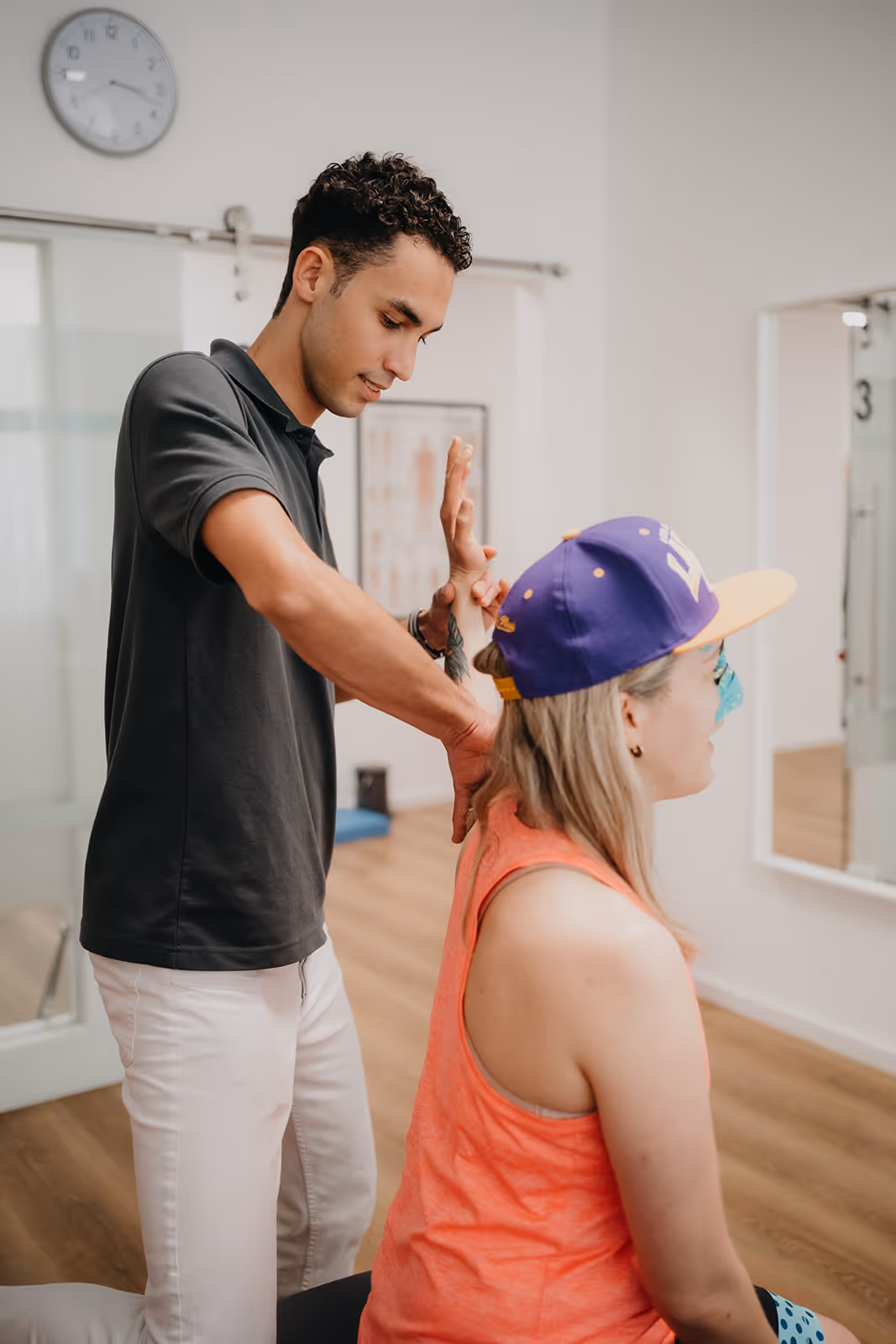 Physiotherapeut untersucht und mobilisiert Arm einer sitzenden Patientin mit lila Baseballkappe in einer Praxis.