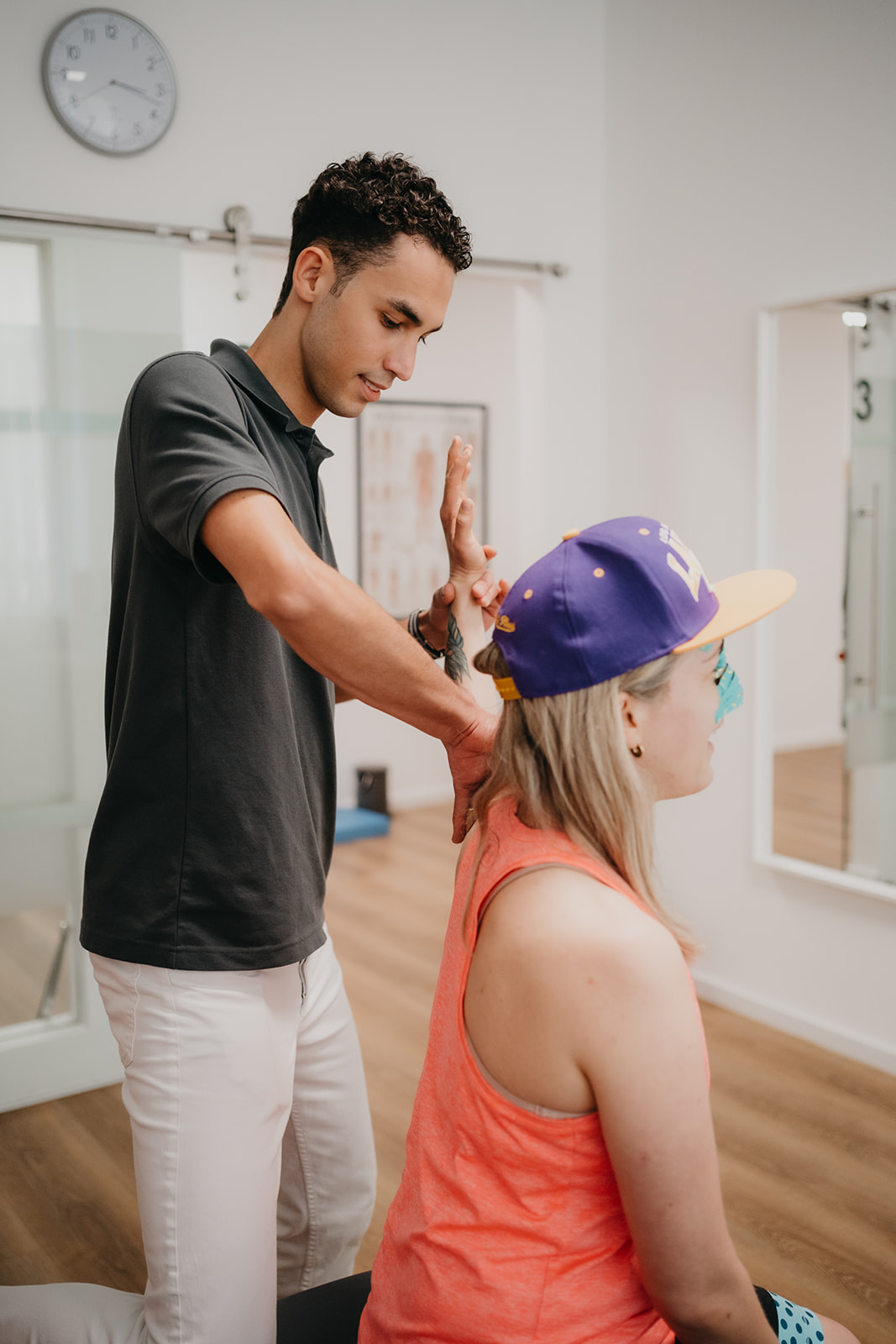 Physiotherapeut untersucht und mobilisiert Arm einer sitzenden Patientin mit lila Baseballkappe in einer Praxis.