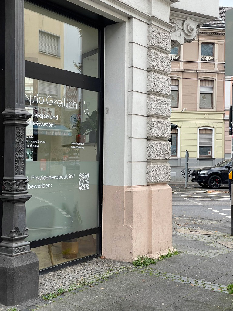 Eingangstür einer Physiotherapiepraxis mit Glasfront und weißen Schriftzügen neben einer historische Gebäudeecke.