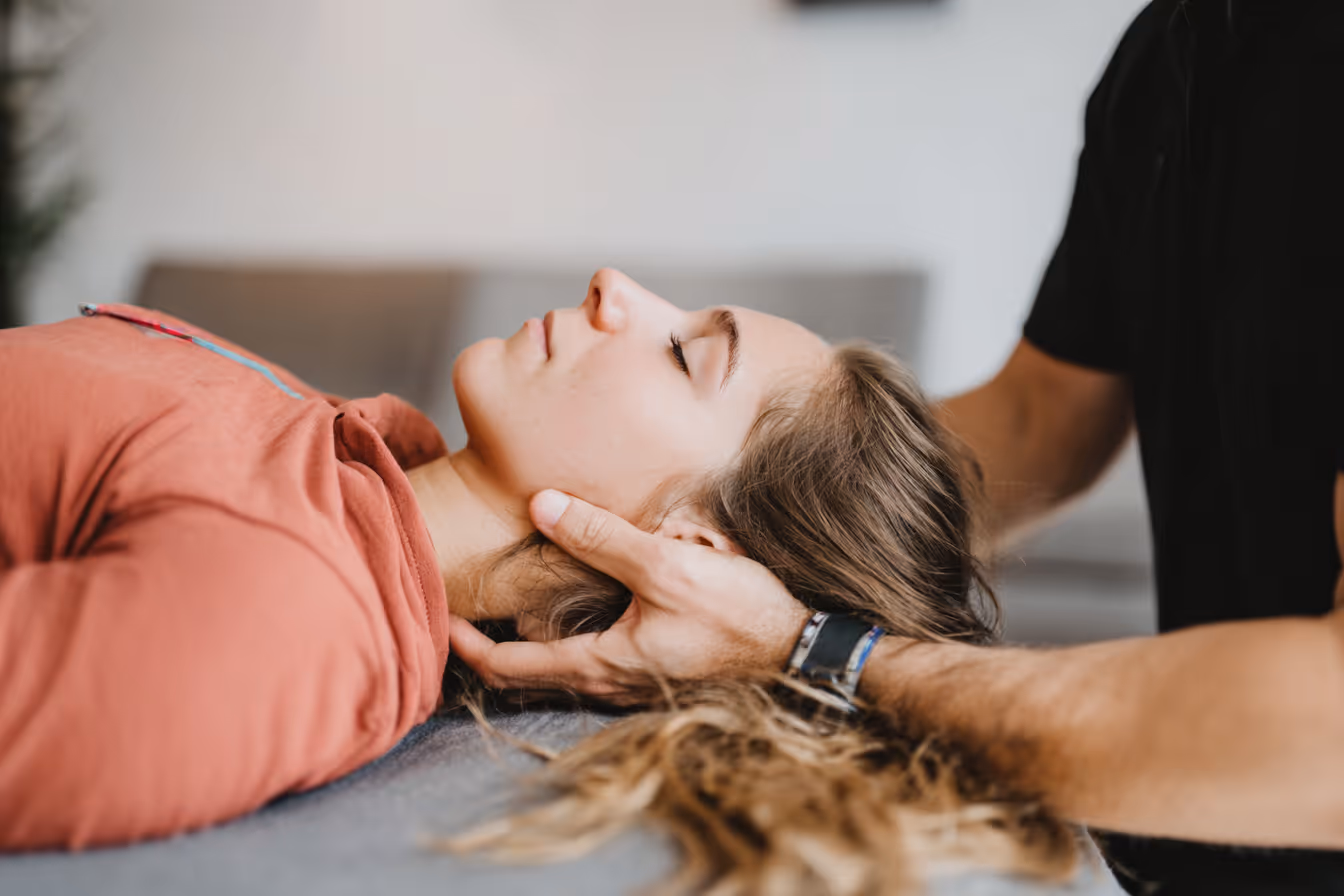 Frau liegt entspannt auf dem Rücken, während eine andere Person ihren Kopf behutsam hält, wahrscheinlich bei einer Physiotherapie.