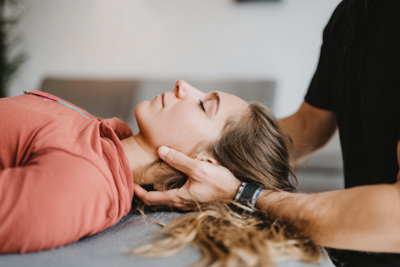 Frau liegt entspannt auf dem Rücken, während eine andere Person ihren Kopf behutsam hält, wahrscheinlich bei einer Physiotherapie.
