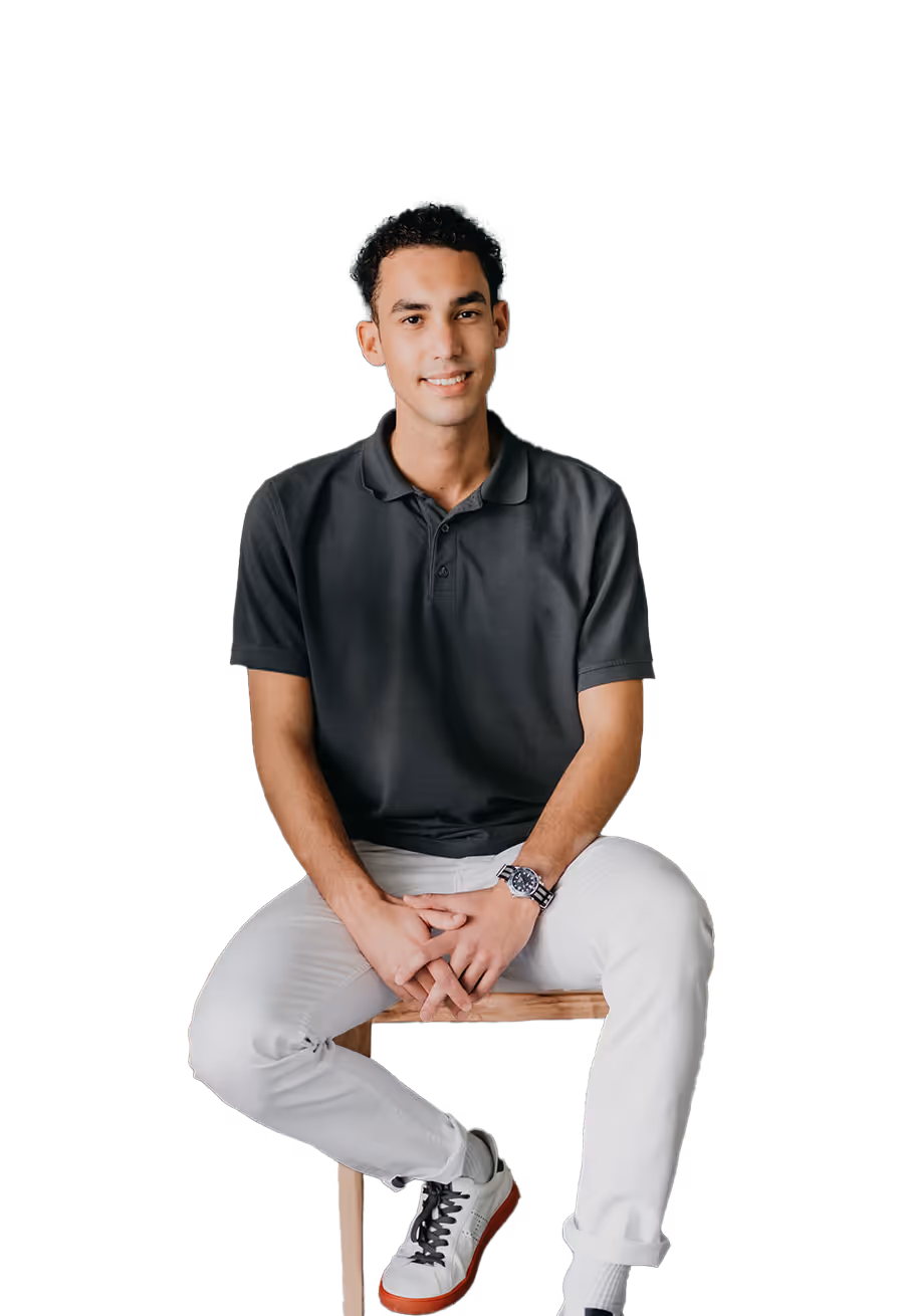 Junger Mann in schwarzem Poloshirt und weißen Hosen sitzt auf einem Holzstuhl, lächelt und verschränkt die Hände.