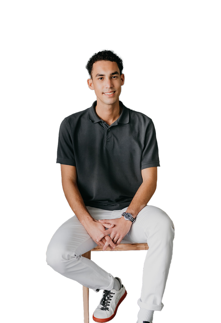 Junger Mann in schwarzem Poloshirt und weißen Hosen sitzt auf einem Holzstuhl, lächelt und verschränkt die Hände.