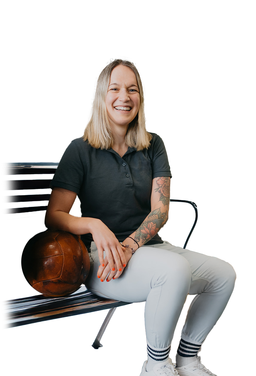 Lächelnde junge Frau mit hellen Haaren und tätowierten Armen sitzt auf einer Bank und lehnt an einem alten braunen Leder-Fußball.