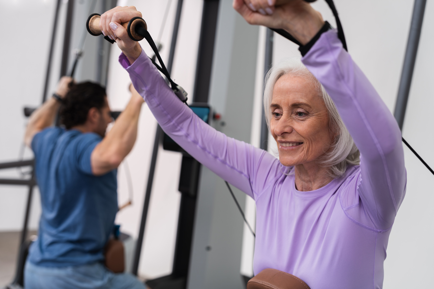 Ältere Frau in lila Sportkleidung trainiert an einem Trainingsgerät, ein Mann im Hintergrund trainiert ebenfalls.