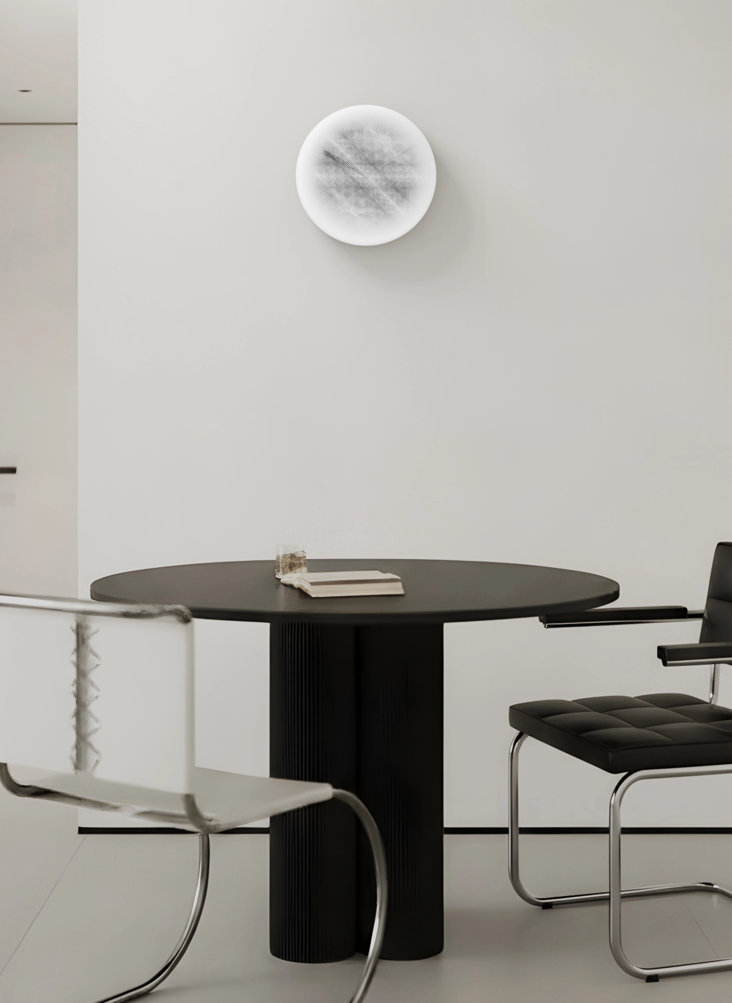 Minimalist dining area with a round black table, a black padded chair, a transparent chair with metal legs, and a white textured circular wall decoration.
