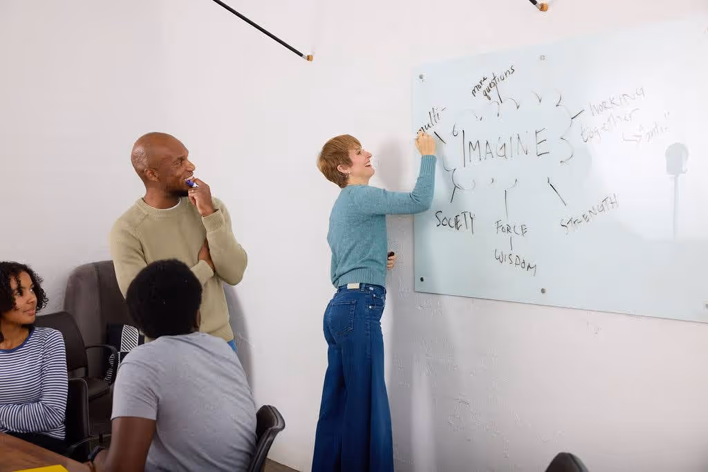 A group of four diverse people collaborating in a meeting room, one writing the word 'IMAGINE' on a whiteboard with related keywords around it.