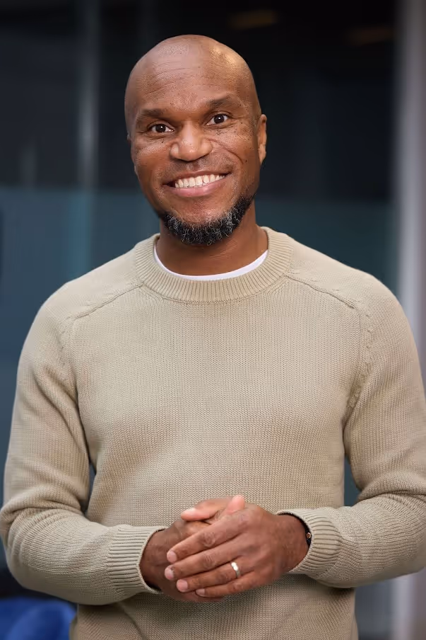 Smiling bald man with a short beard wearing a beige sweater, standing with hands clasped indoors.