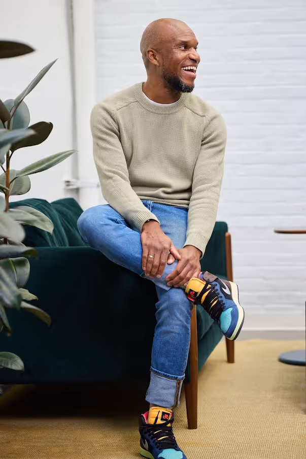 Smiling man with beard sitting cross-legged on a dark green sofa wearing a beige sweater, blue jeans, and colorful sneakers.