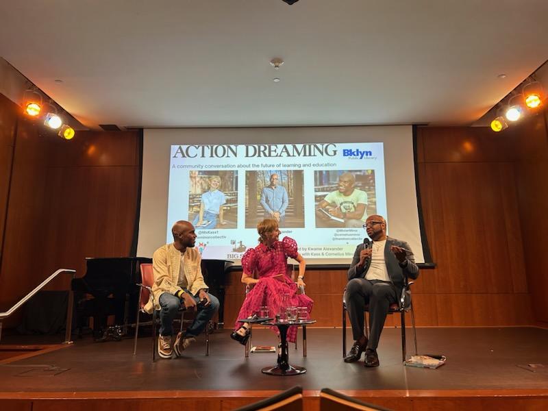 Three people seated on a stage engaged in a discussion under a screen displaying 'Action Dreaming' event about learning and education.