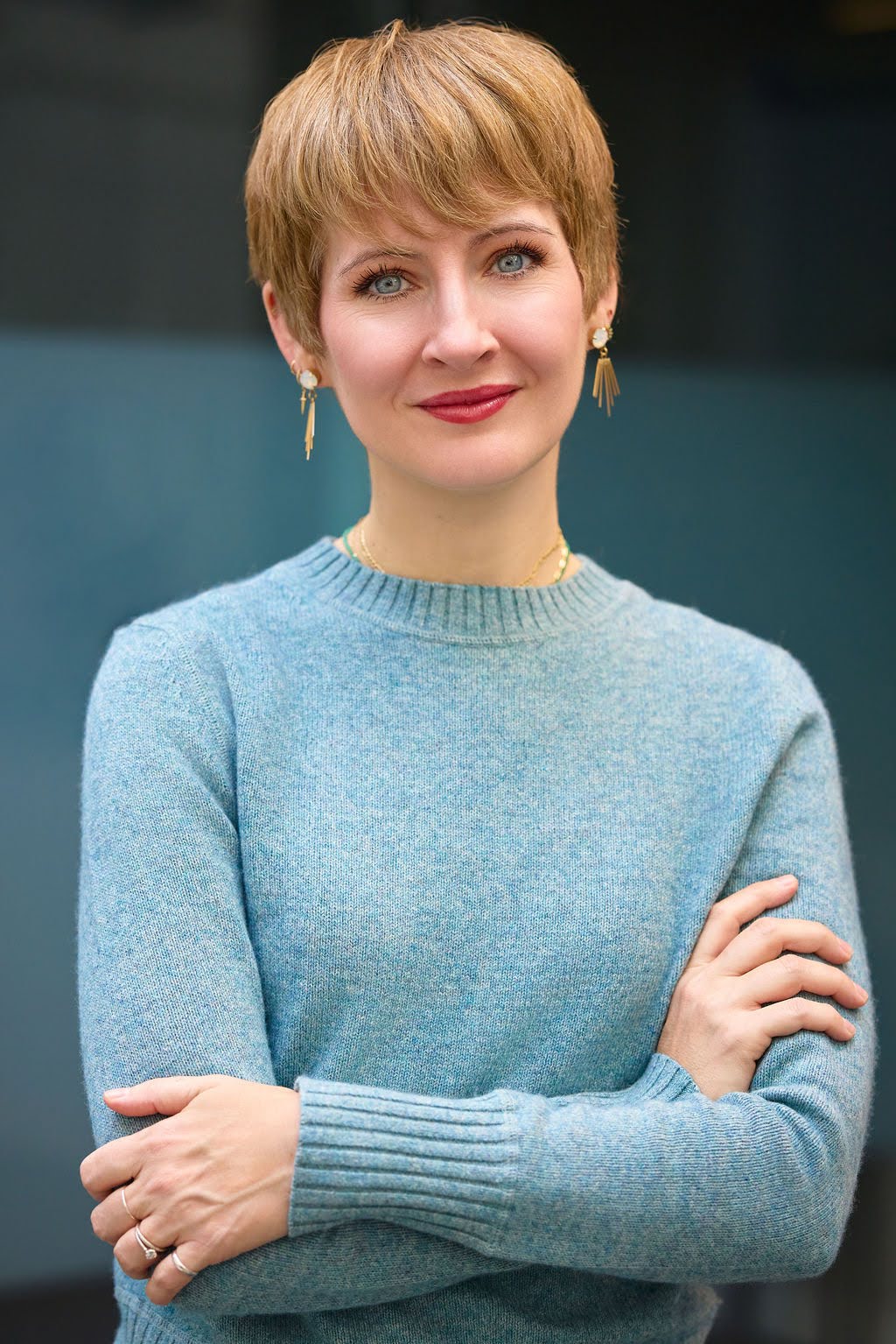 Portrait of a woman with short light brown hair, wearing a blue sweater and gold earrings, standing with arms crossed.